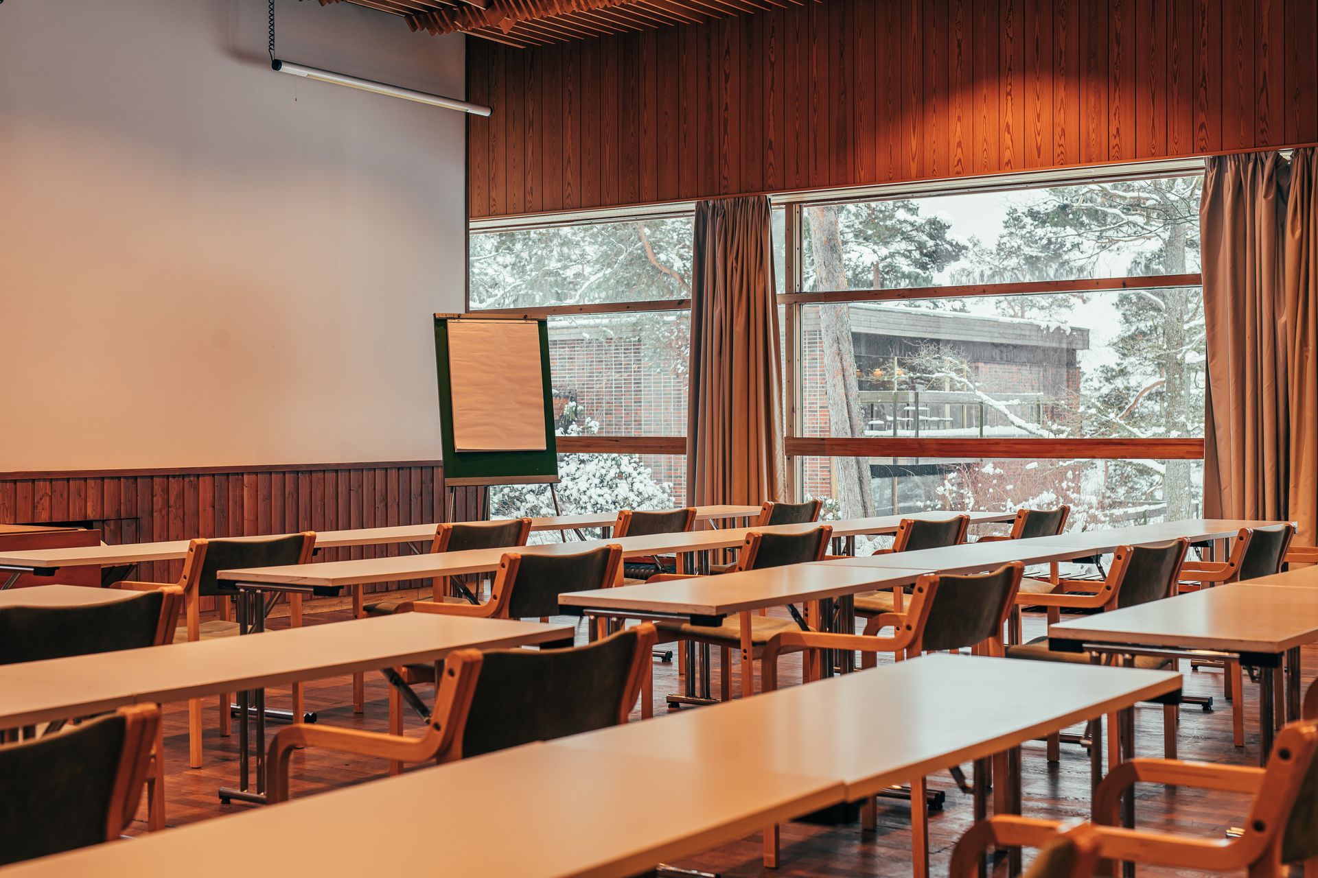 Tyhjä kokous- tai neuvotteluhuone, jossa on rivit puisia pöytiä, tuoleja ja suuria ikkunoita, joista on näkymät lumiseen metsään.