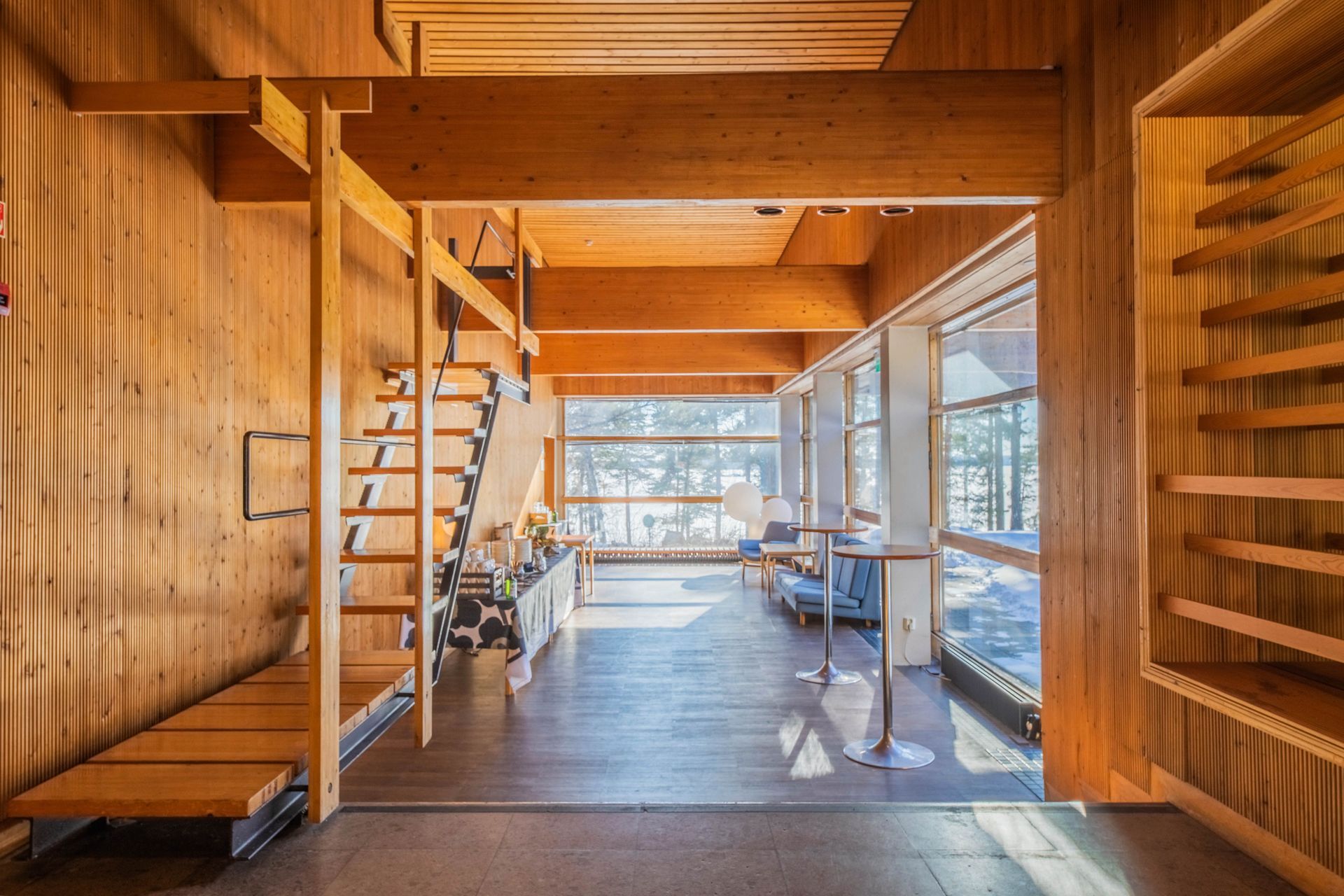 Maalaismainen huone, jossa on lämpimät puuseinät, ullakkoportaat, avohyllyt ja suuret ikkunat, joista on näkymät puihin.