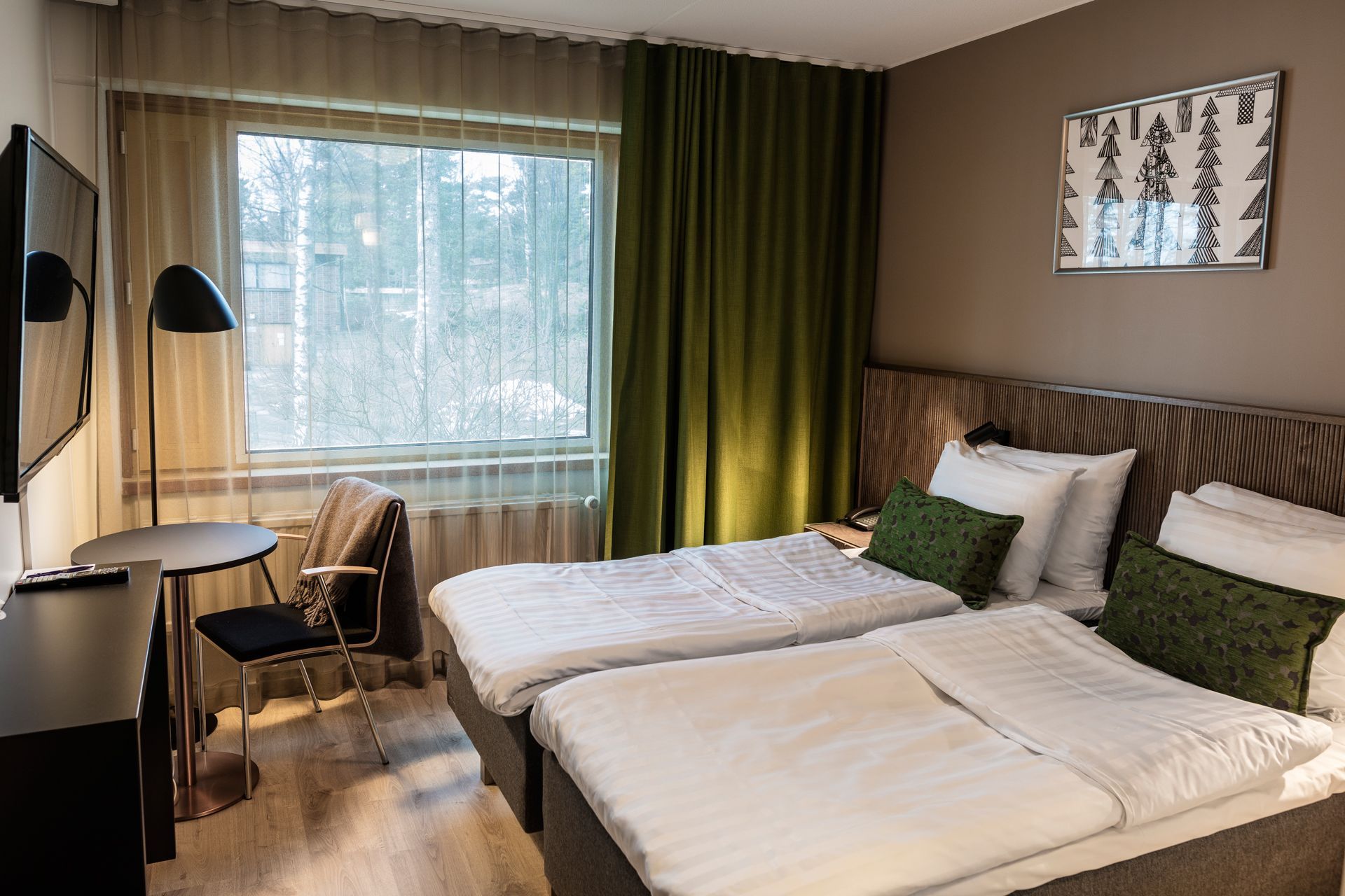Hotellihuone, jossa on kaksi erillistä sänkyä vihreillä koristetyynyillä, pieni työpöytä, tuoli ja ikkuna vihreillä verhoilla.