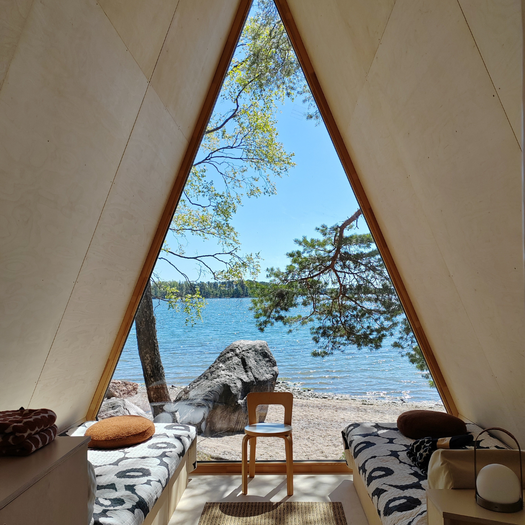 Kolmionmuotoinen puinen mökkirunko kehystää näkymän järvelle ja kallioiselle rannalle, ja sen sisällä on kaksi sänkyä ja tuoli.