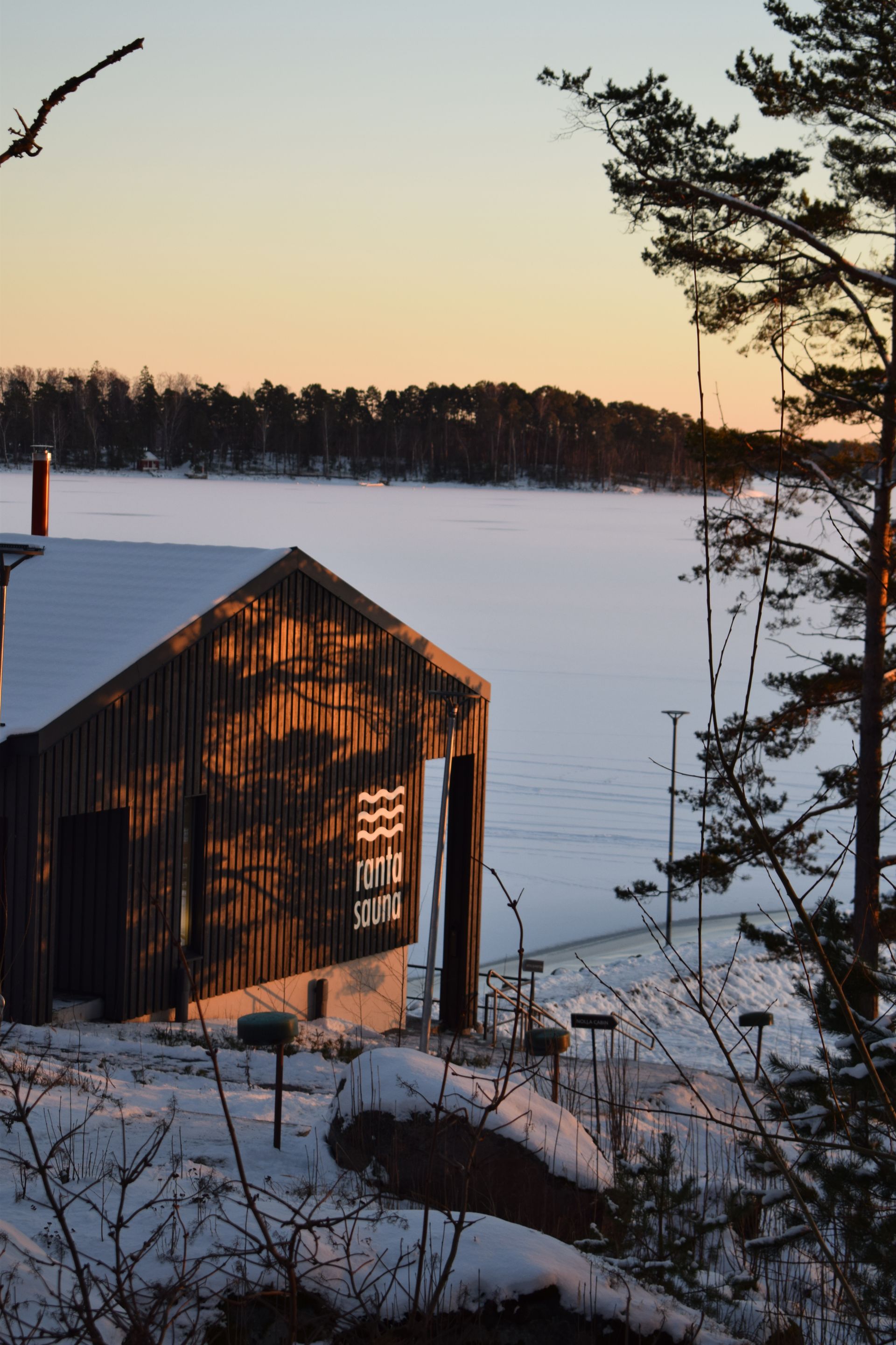 Puinen saunarakennus lumisella, jäätyneellä järvenrannalla auringonlaskun aikaan, mäntyjen reunustamana.