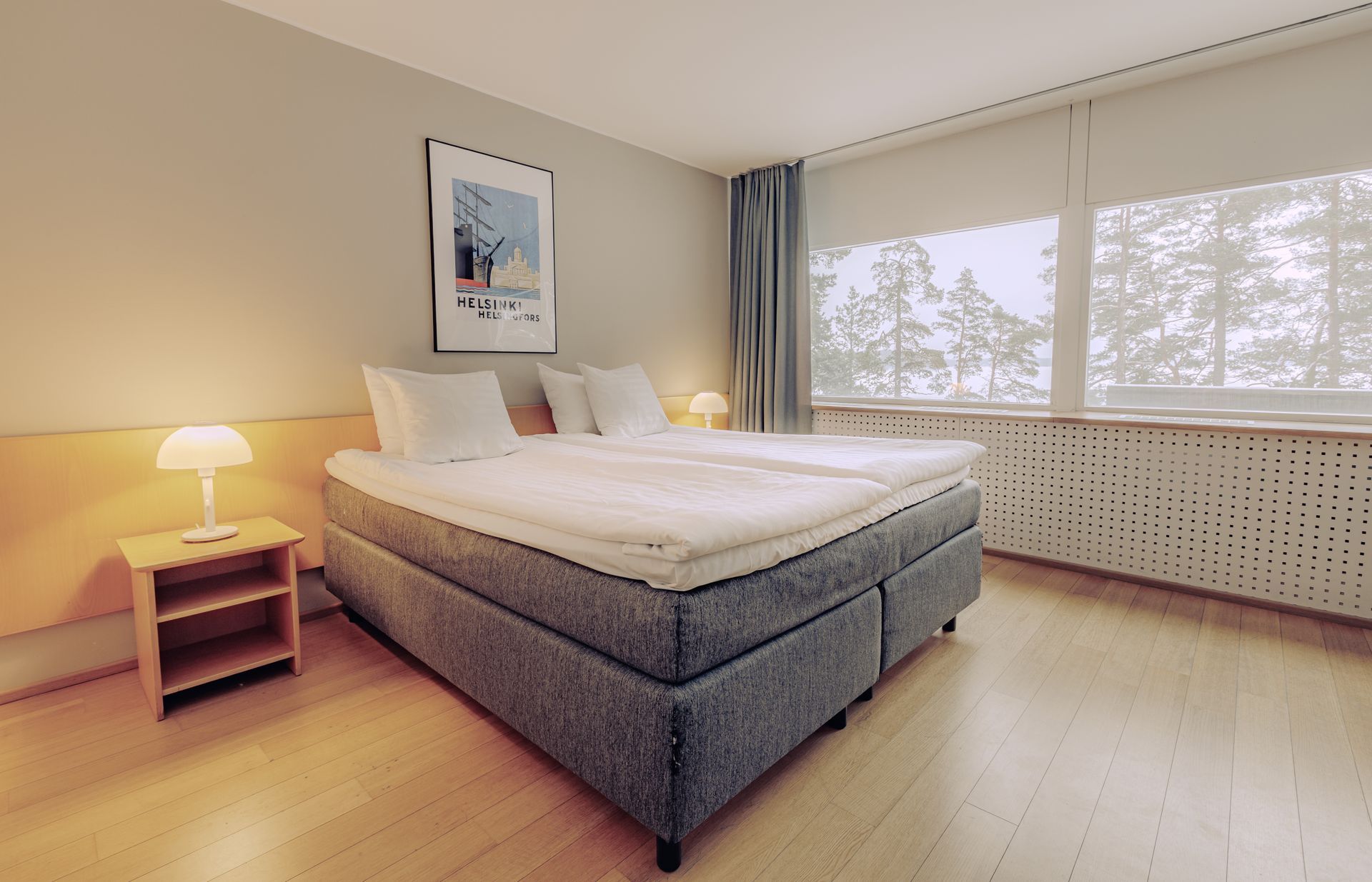 A modern hotel bedroom with a queen bed, wooden nightstand, and large window overlooking a forest.