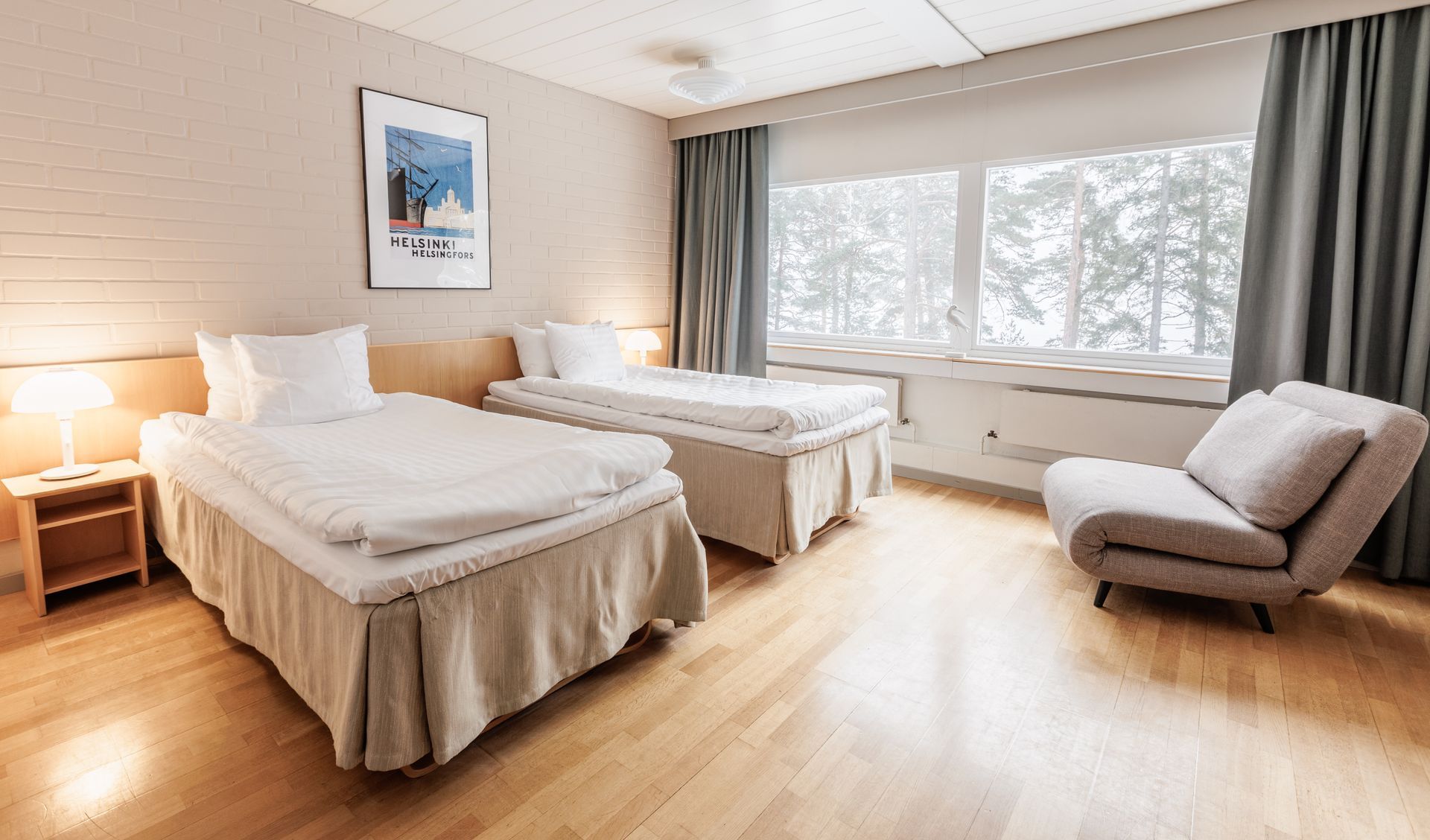 A hotel room with two twin beds, a small armchair, a nightstand, and a large window overlooking a snowy forest.