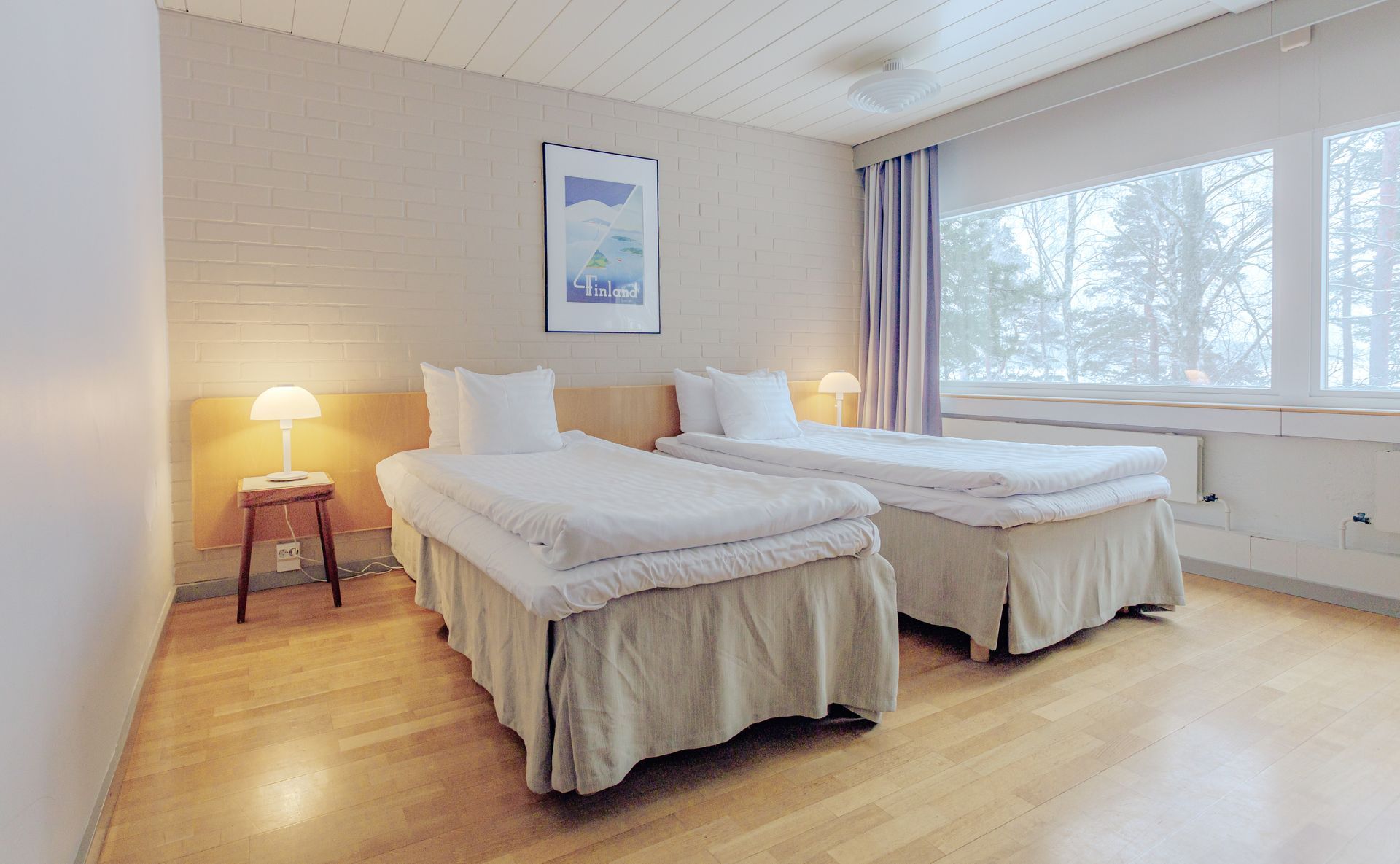 Hotellihuone, jossa on kaksi erillistä sänkyä valkoisine liinavaatteineen, vaaleat puulattiat ja yöpöytälamppu ikkunan vieressä.