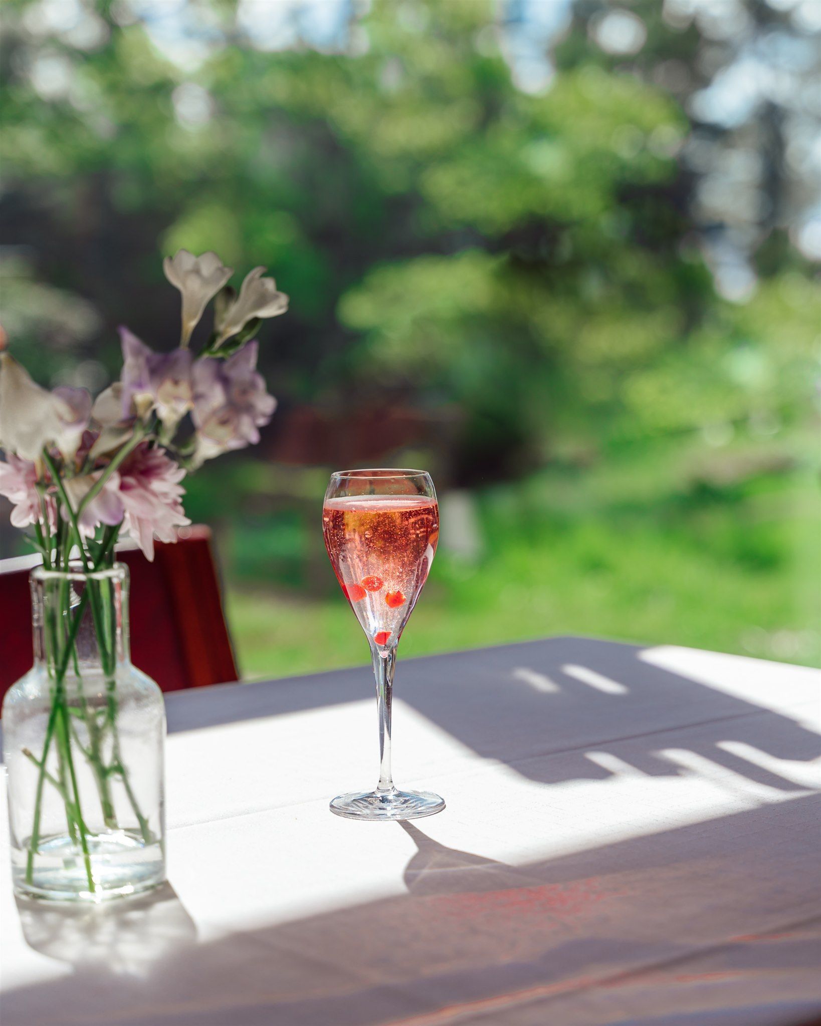 A glass of rose-colored sparkling drink with berries and a vase of flowers on a white table outdoors.