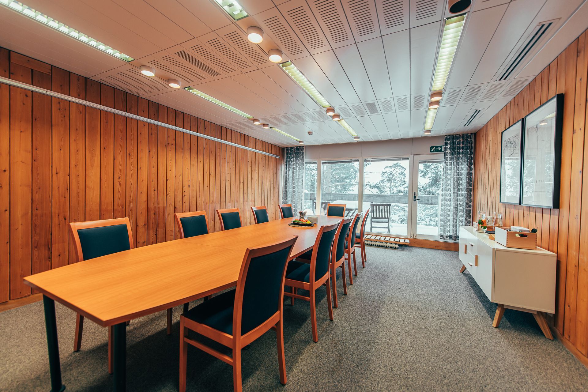 Kokoushuone, jossa on pitkä puupöytä ja tummat tuolit, puupaneloidut seinät, valkoinen sohva ja suuret ikkunat.