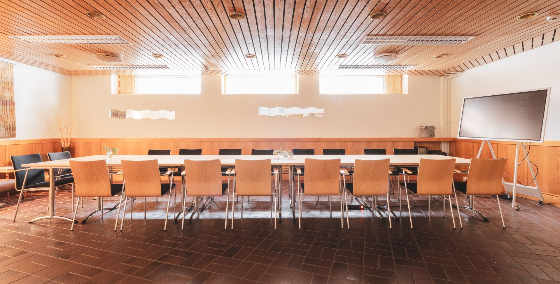 Pitkä puinen neuvottelupöytä tuoleineen huoneessa, jossa on sälepohjainen puukatto ja valkoinen kuivapyyhittävä taulu.