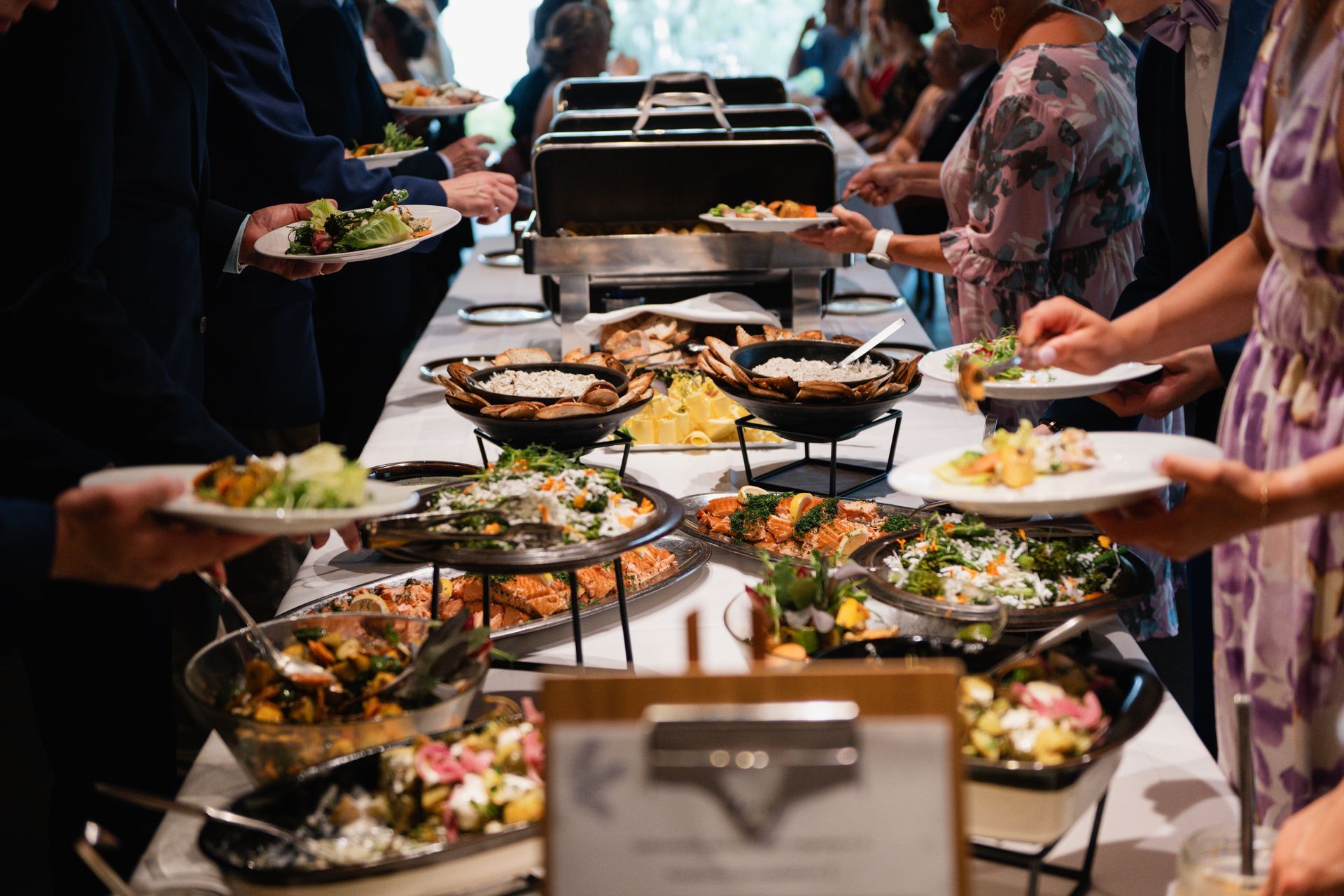 Buffet-jono, jossa ihmiset täyttävät lautasensa erilaisilla annoksilla useista tarjoilukulhoista ja kuumailmakulhoista.