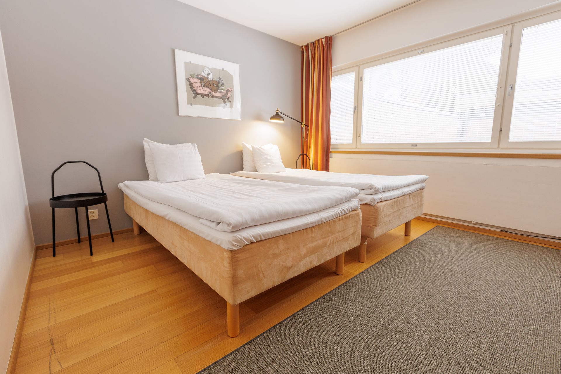 Two single beds with white bedding in a minimalist bedroom with wood floors, a grey wall, and a large window.