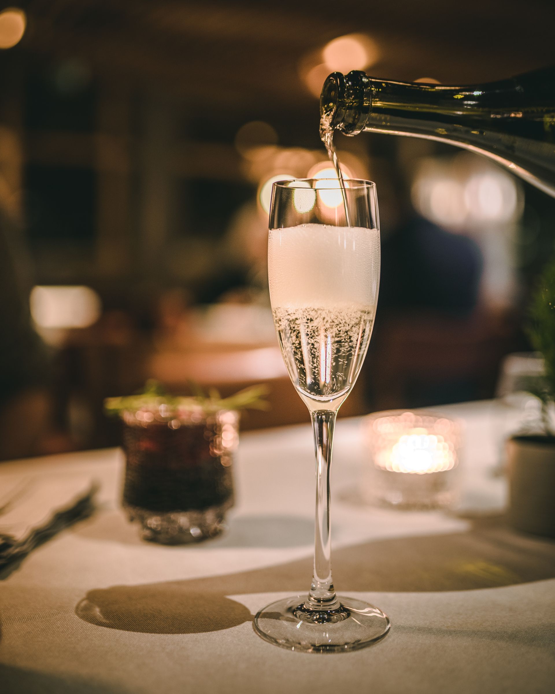 A glass of champagne being poured from a bottle at a softly lit table in a restaurant.