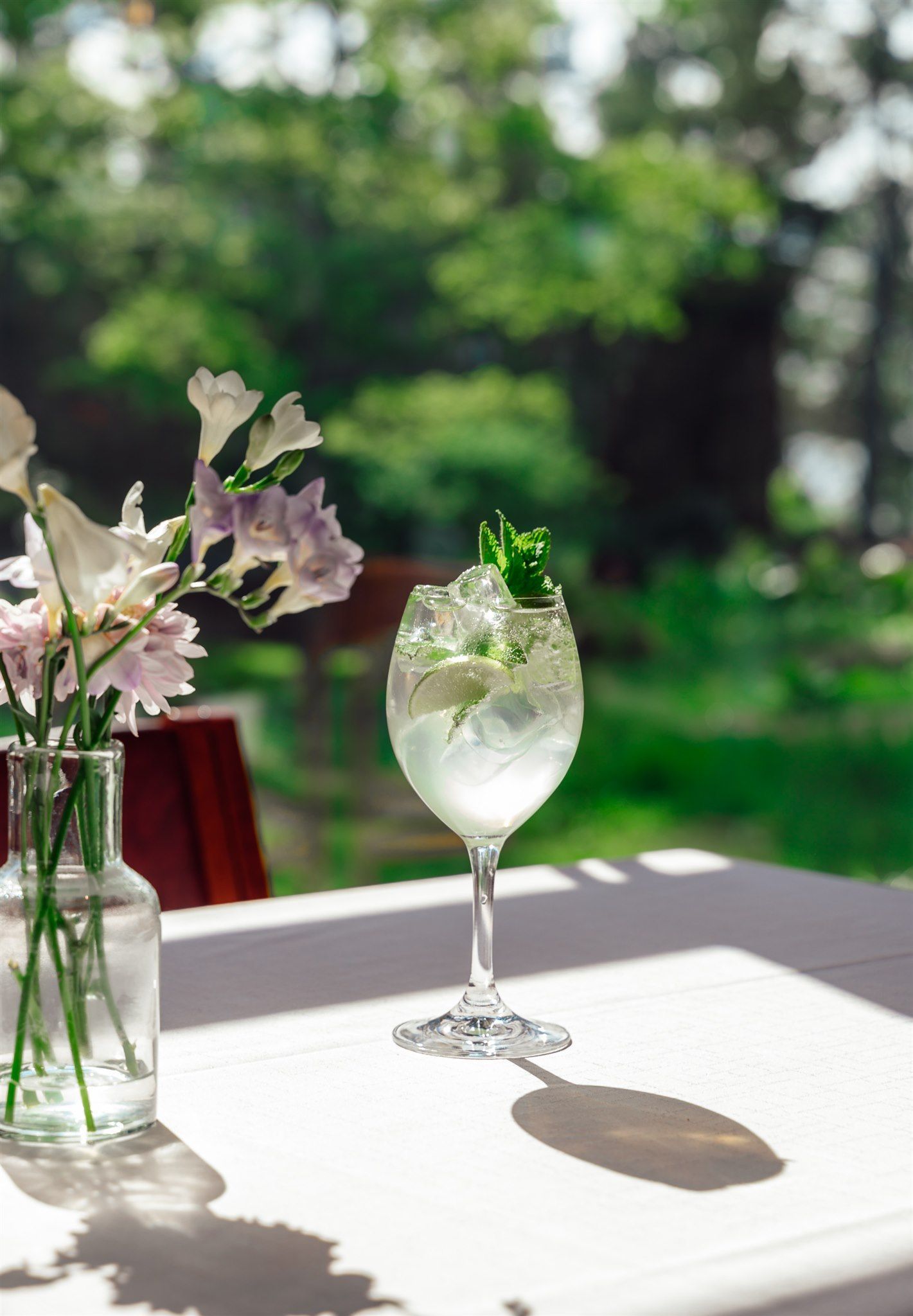 Lasillinen jääcocktailia minttukoristeella vaaleanväristen kukkien maljakon vieressä pöydällä aurinkoisessa puutarhassa.