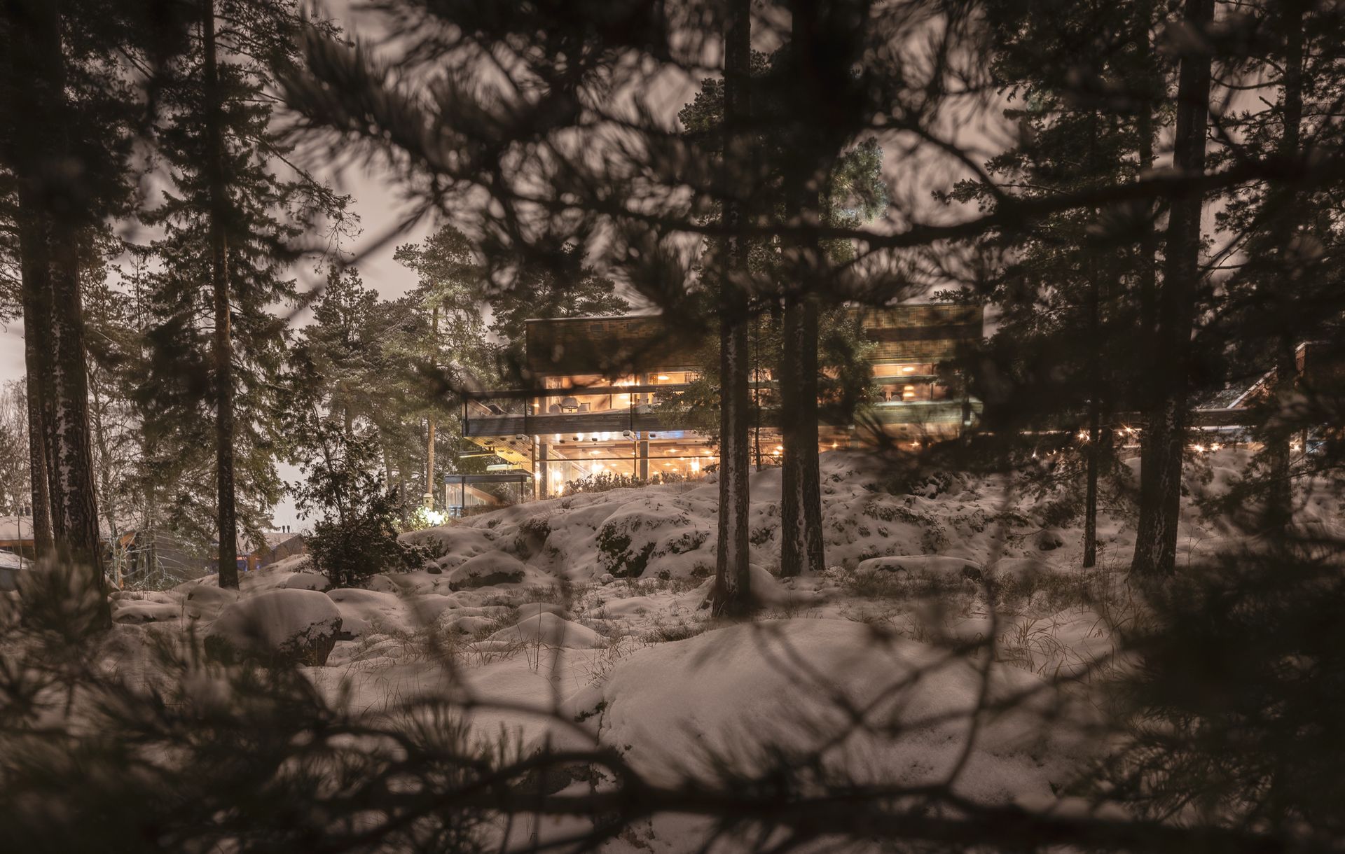 A brightly lit cabin glows through a dark, snow-covered forest at night.