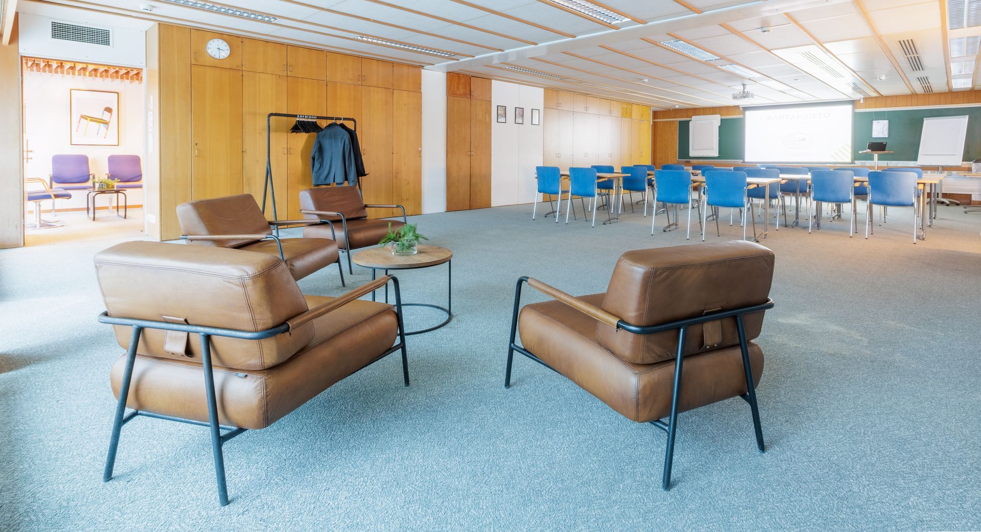 Etualalla oleskelutila, jossa on ruskeita nahkatuoleja ja pyöreä pöytä, ja taustalla kokoushuone.