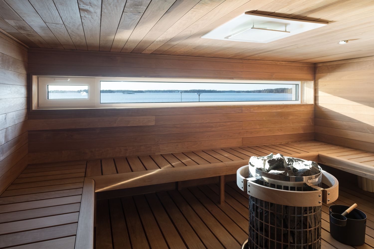 Setripuinen sauna, jossa on porrastetut penkit ja kivinen kiuas, ja vaakasuora ikkuna, josta on näkymä vesistöön.
