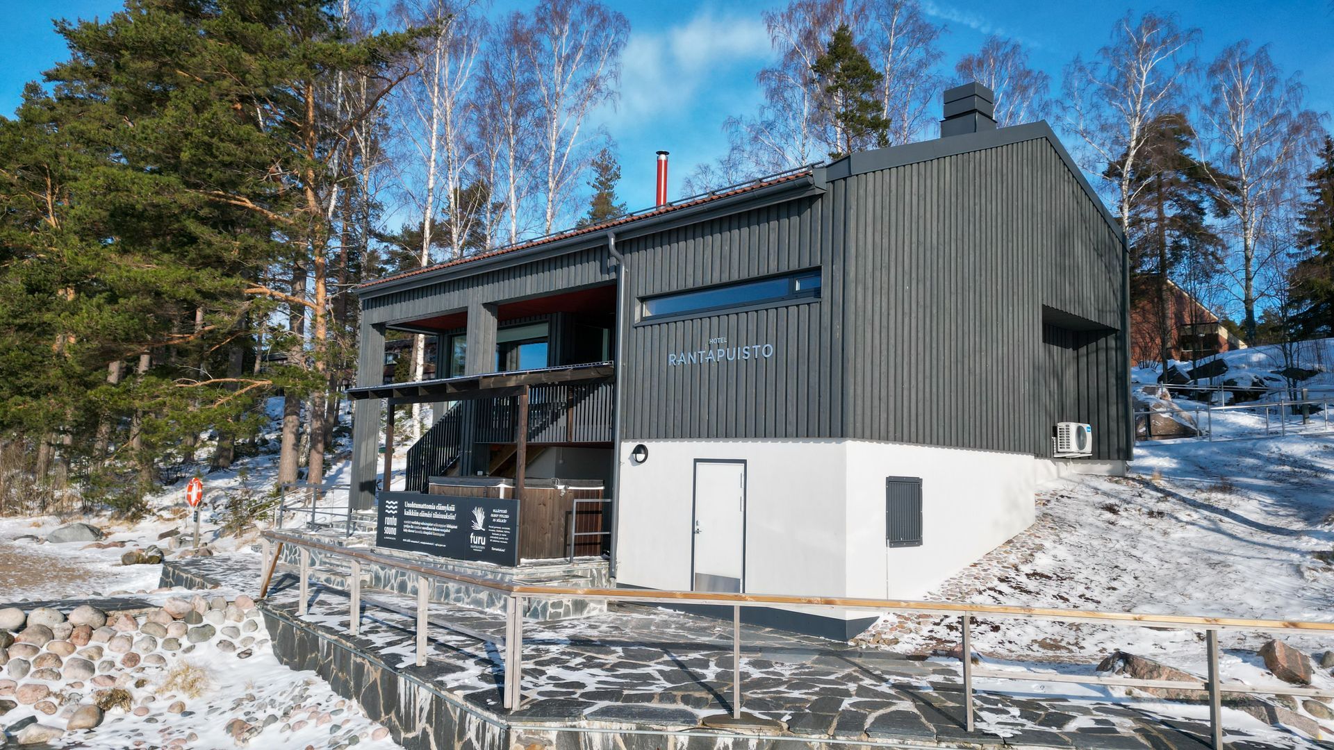 Moderni, kaksikerroksinen mökki, jossa on harmaa puuverhous ja valkoinen alakerta, lumimaisemassa.