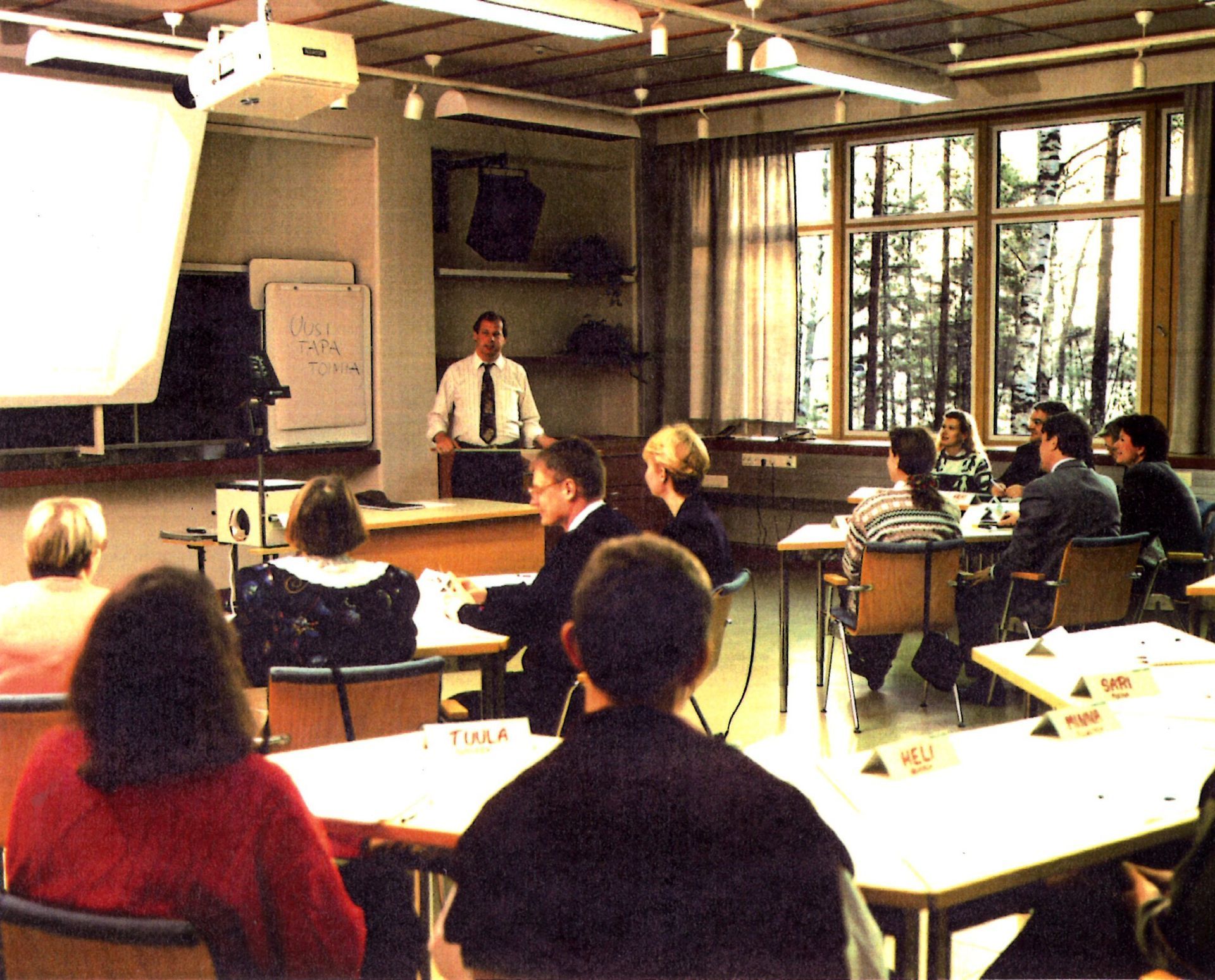 Esittäjä seisoo pienen ihmisryhmän edessä pöydissä kirkkaasti valaistussa luokkahuoneessa, jossa on suuret ikkunat.