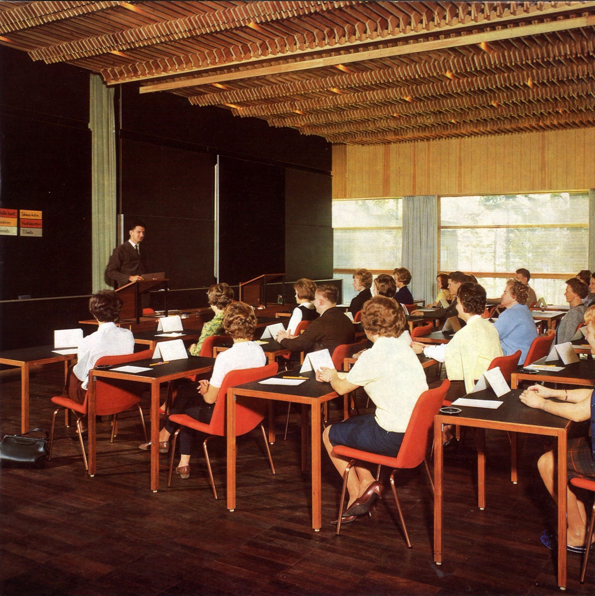 Puhuja seisoo yleisön edessä pöytien ääressä kokoushuoneessa, jossa on puinen sälekatto ja punaiset tuolit.