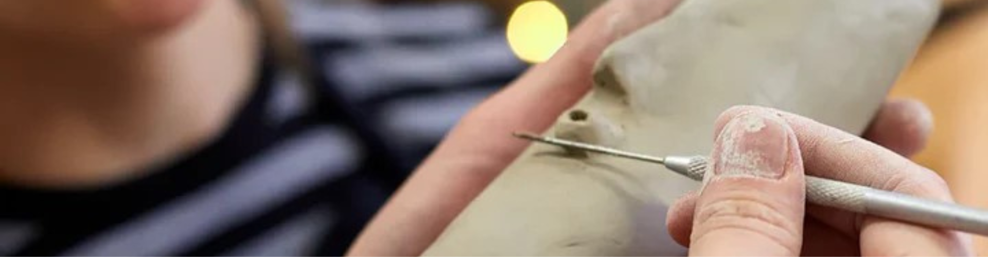 Lähikuva käsistä, jotka käyttävät metallista veistotyökalua kaivertaakseen hienoja yksityiskohtia harmaaseen saveen.