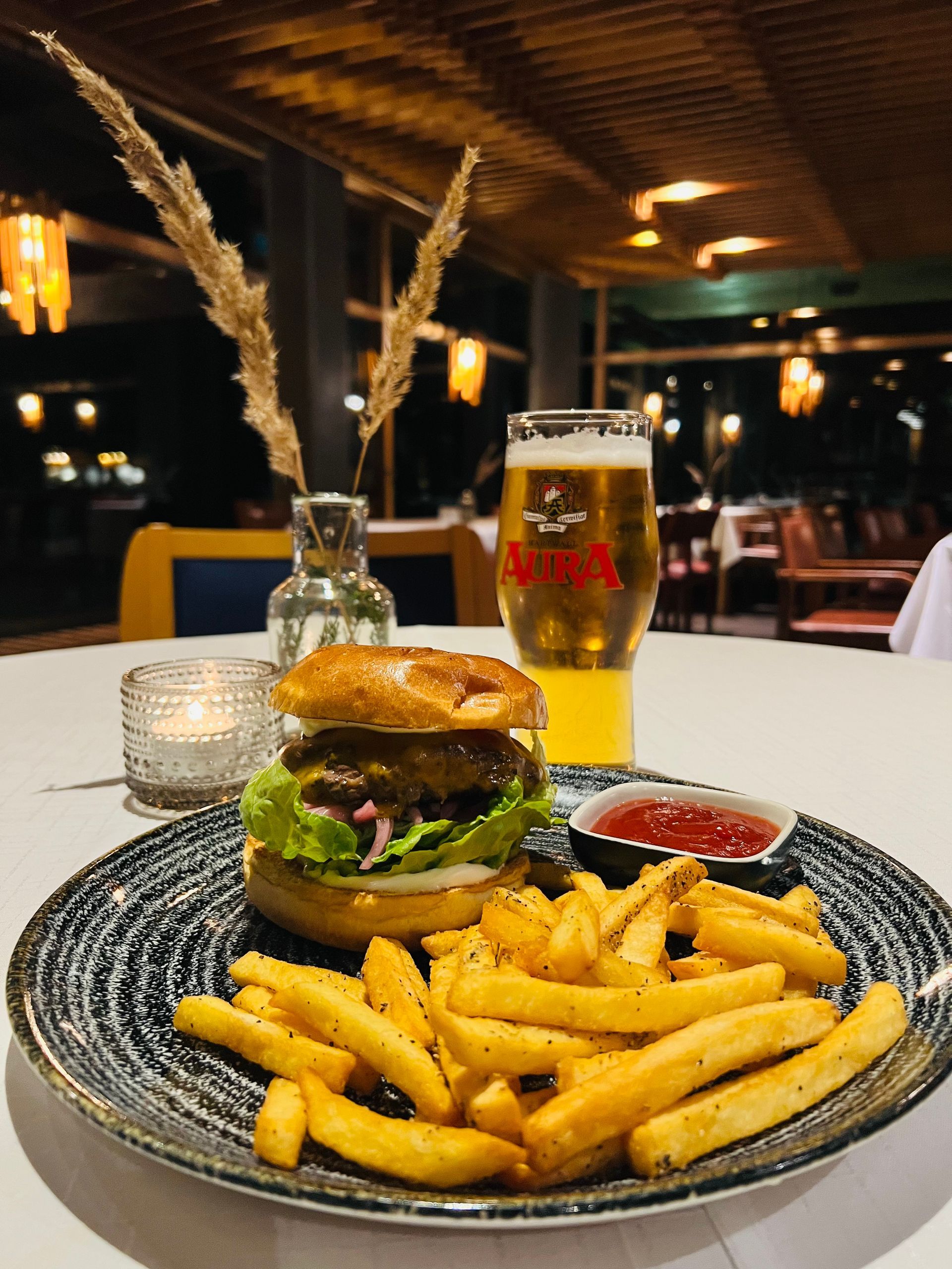 Hampurilainen ranskalaisten, ketsupin ja lasillisen olutta täplikkäällä lautasella ravintolaympäristössä.