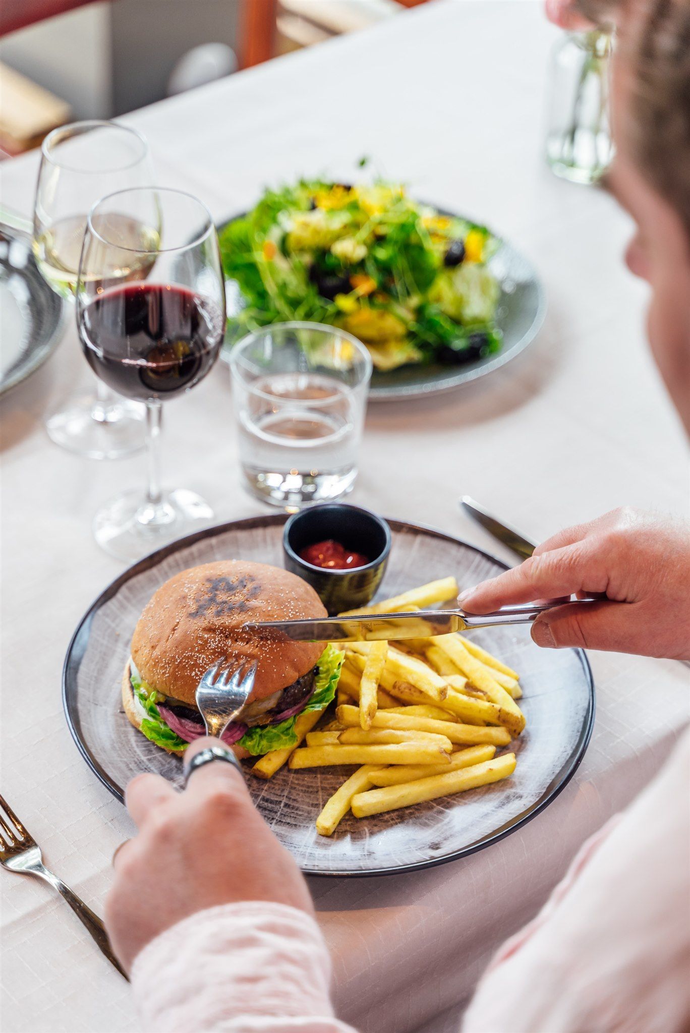 Henkilö leikkaa hampurilaisen lautaselle ranskalaisten perunoiden kera, lisukesalaatin ja kahden lasillisen viiniä kanssa.