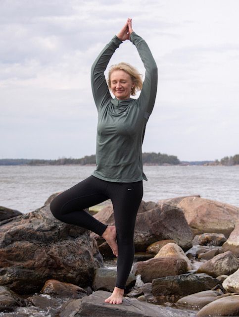 Henkilö harjoittelee puuasentoa urheiluvaatteissa kallioisella rannalla vesistön taustalla.