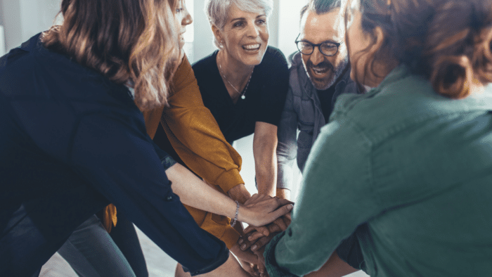 Monimuotoinen ihmisryhmä seisoo piirissä kädet pinossa keskellä, hymyilevät ja katsovat alas.