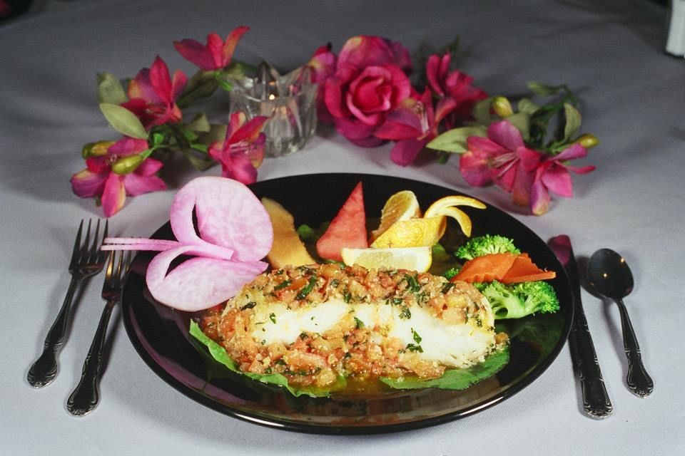 Plate of breaded fish with fruit and vegetables, arranged on a table with silverware and flowers.