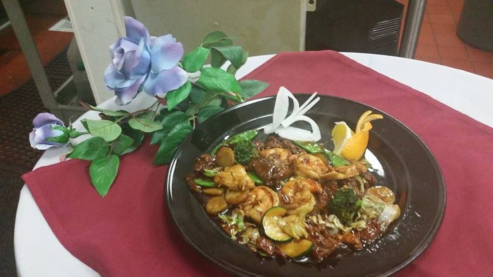 Plate of food with shrimp and vegetables on a table, garnished with onion and orange slice, near a blue flower.