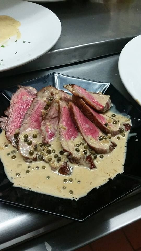 Sliced steak with peppercorn sauce on a black plate, with other plates in the background.