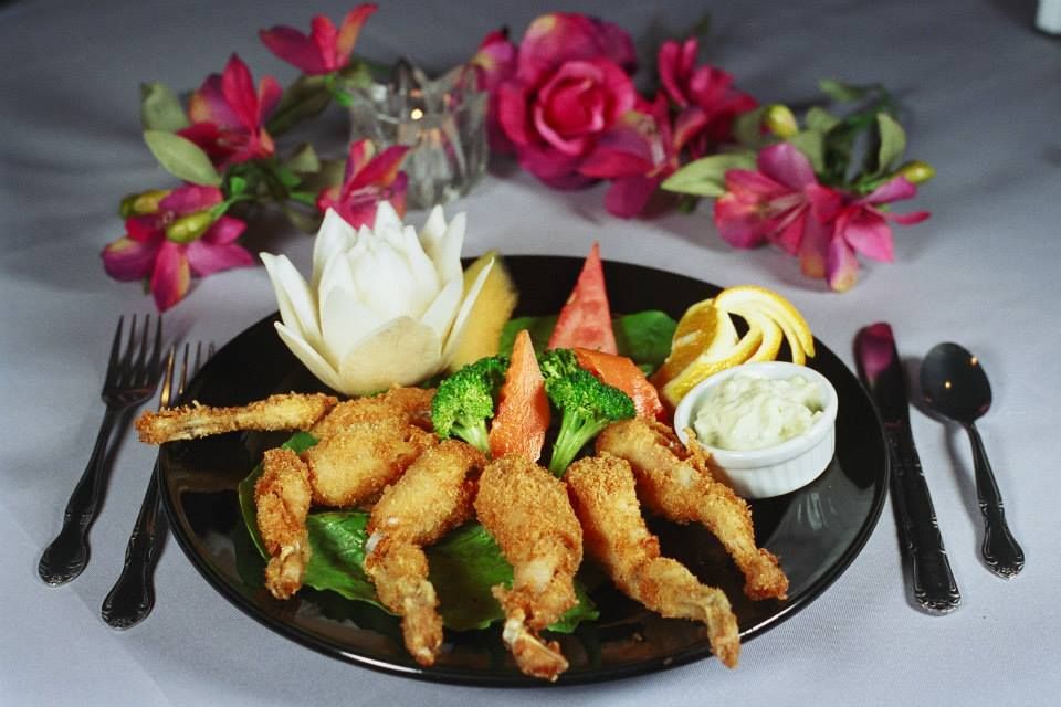 Fried frog legs on a black plate with vegetables, lemon, and a sauce, set on a white tablecloth.