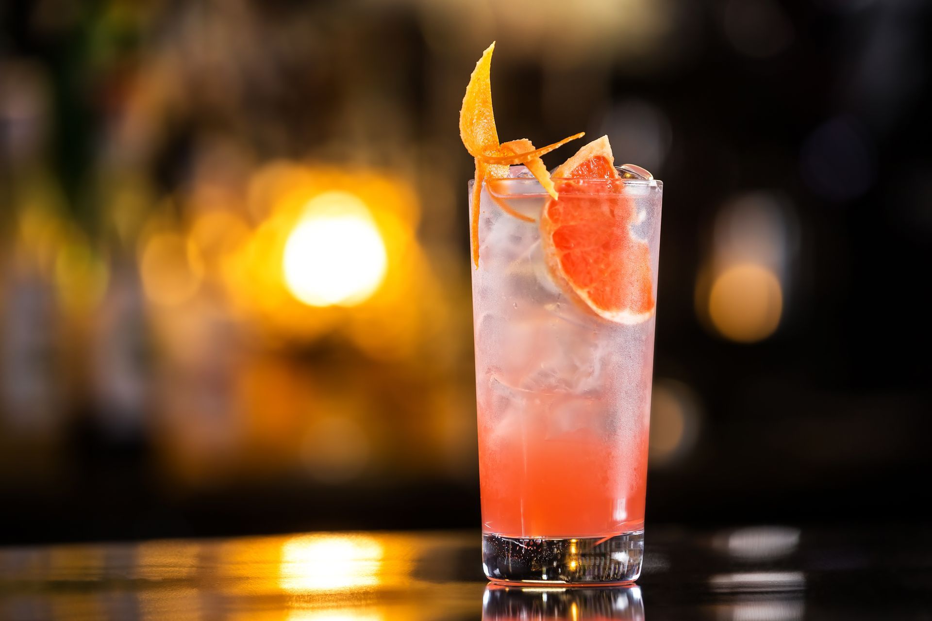 Tall cocktail with a grapefruit slice and peel garnish; set on a bar with a blurred background.