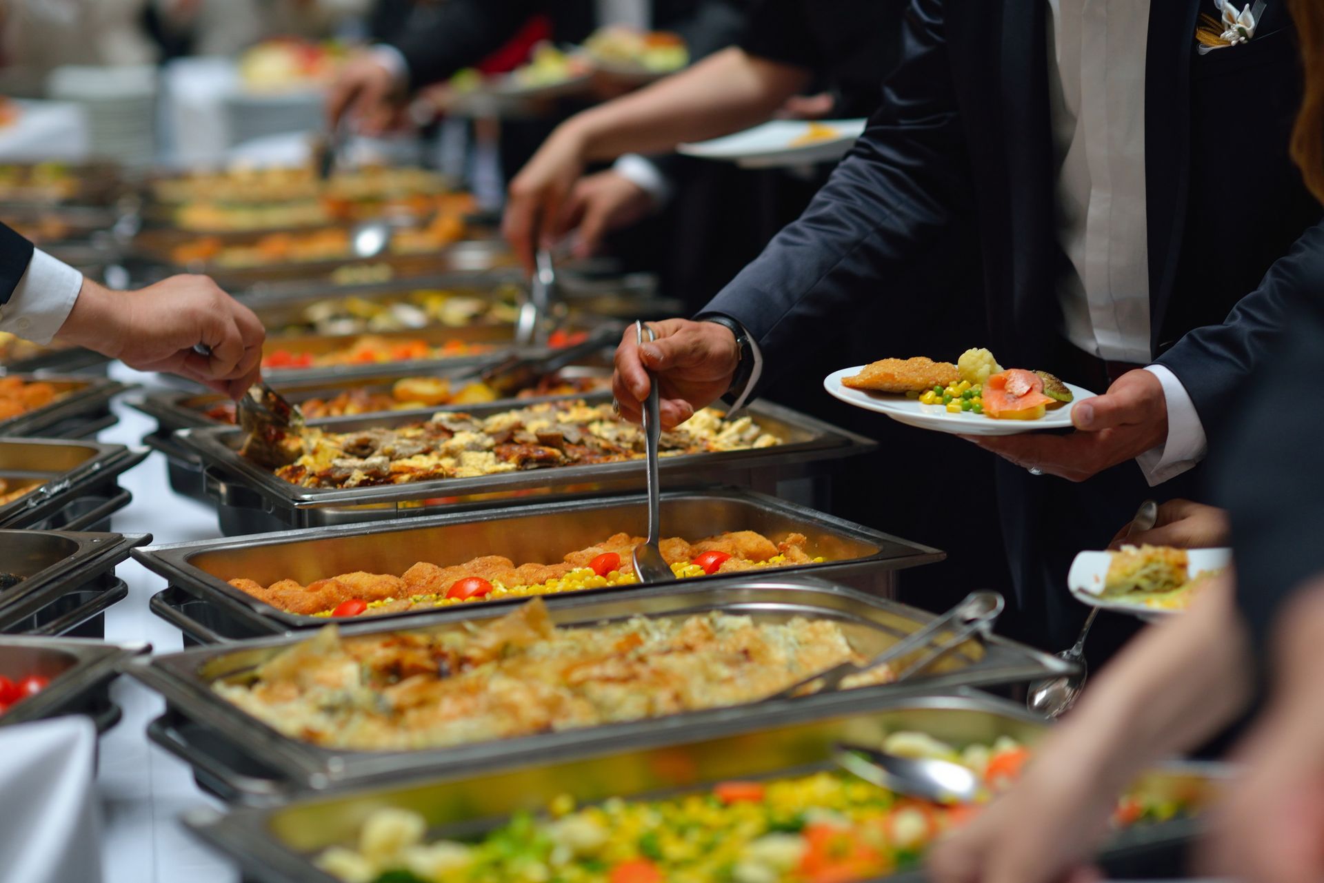 People at a buffet serving themselves various dishes from stainless steel trays.