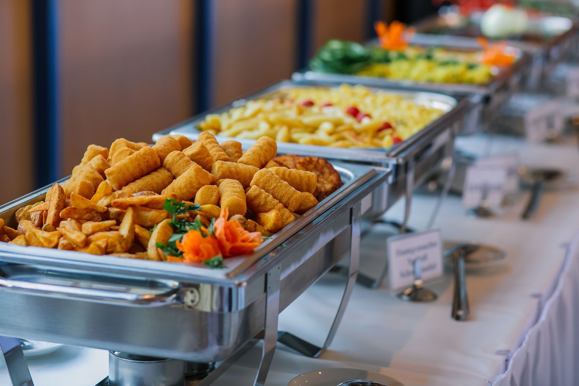 Buffet table with various food items in metal chafing dishes on a white tablecloth.