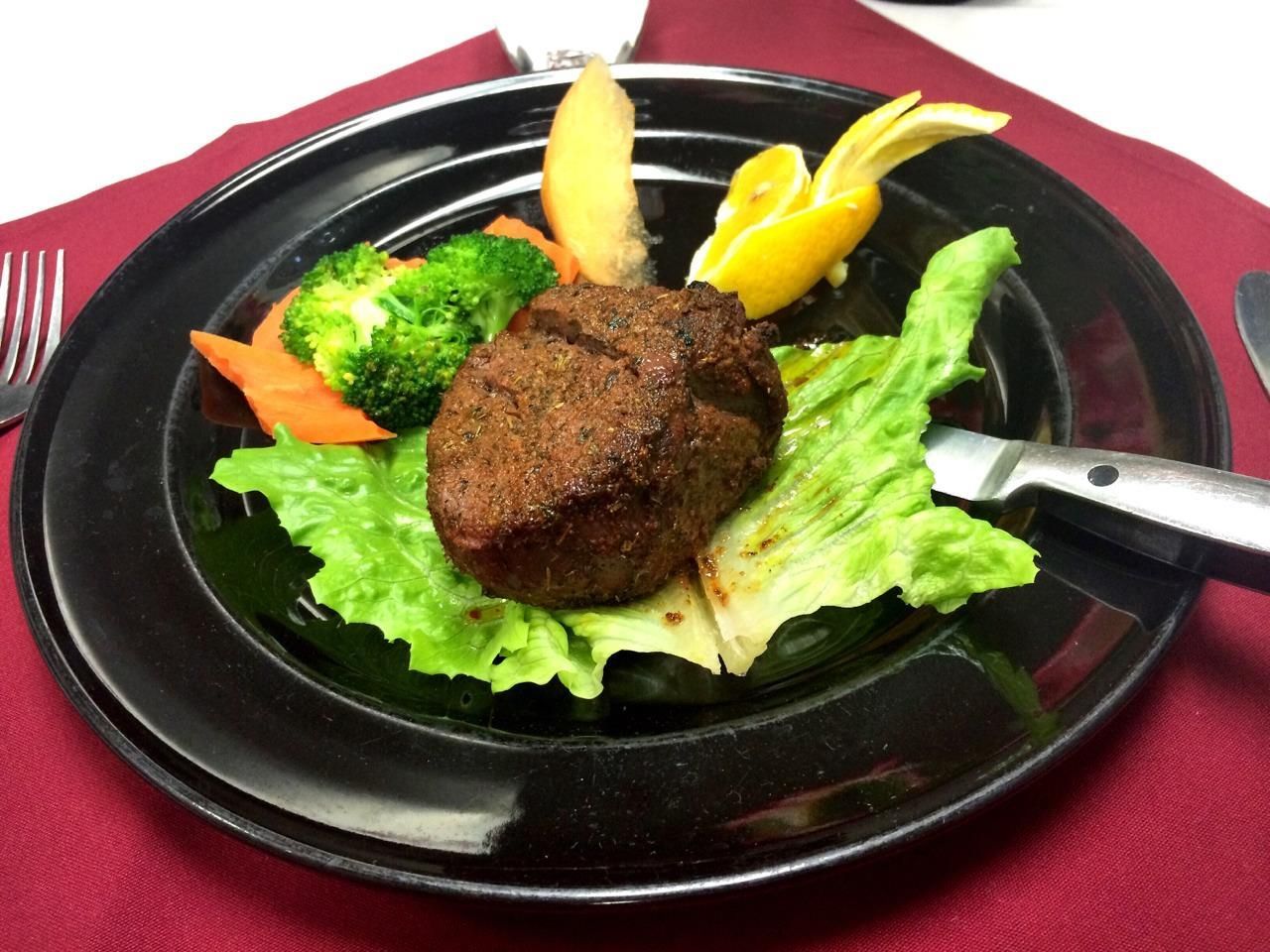 Steak dinner on black plate: seared meat, lettuce, broccoli, carrot, lemon, and knife on red tablecloth.