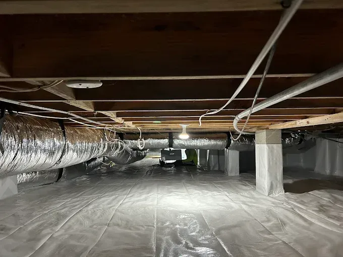 View of a crawl space with white plastic ground covering, HVAC ducting, support posts, and exposed wooden beams.