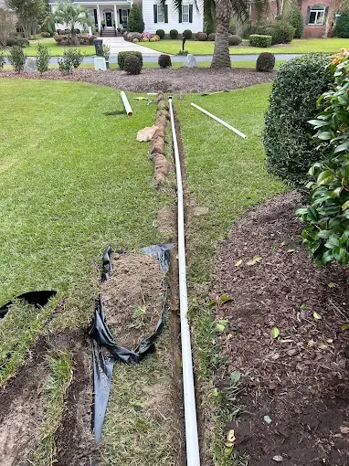 A trench dug in a grassy yard with white pipe laid, for a drainage project.