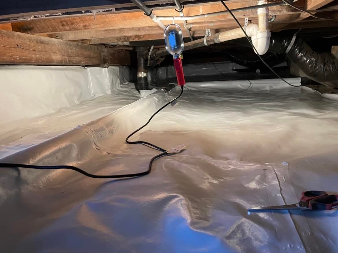 Crawl space with white vapor barrier, plumbing, and a power tool hanging from above.