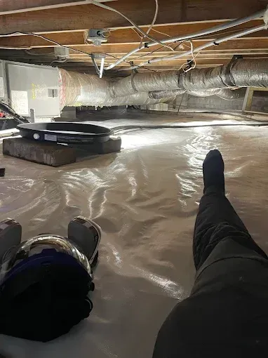 Person lying on a white plastic vapor barrier in a crawl space. Legs and boots visible.