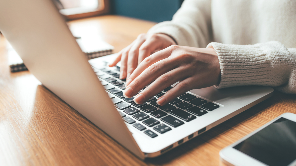 Person typing on a laptop at a wooden desk, next to a phone.