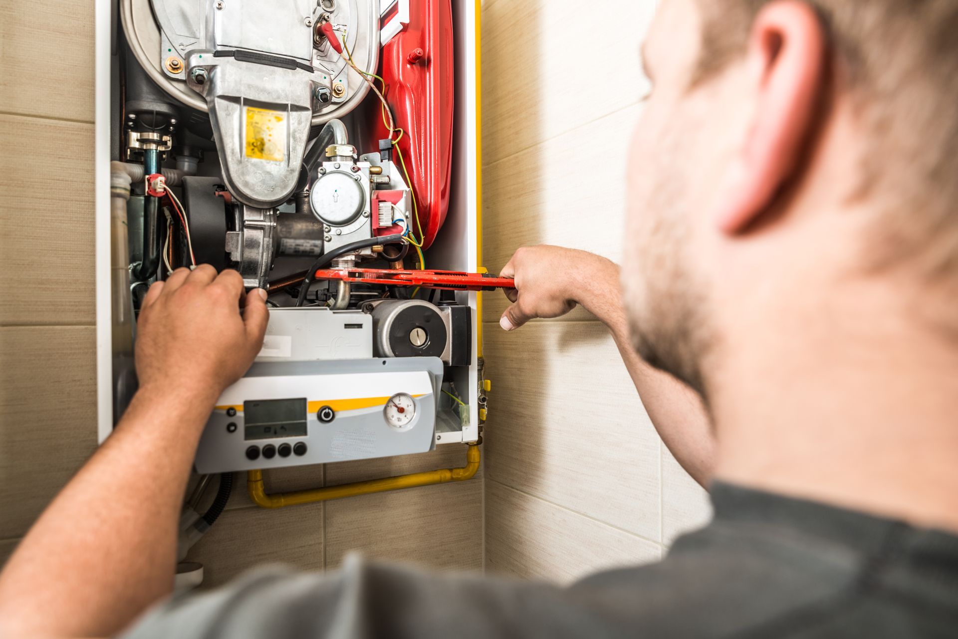 A man repairs a white and red boiler with a wrench. He is inside a building.