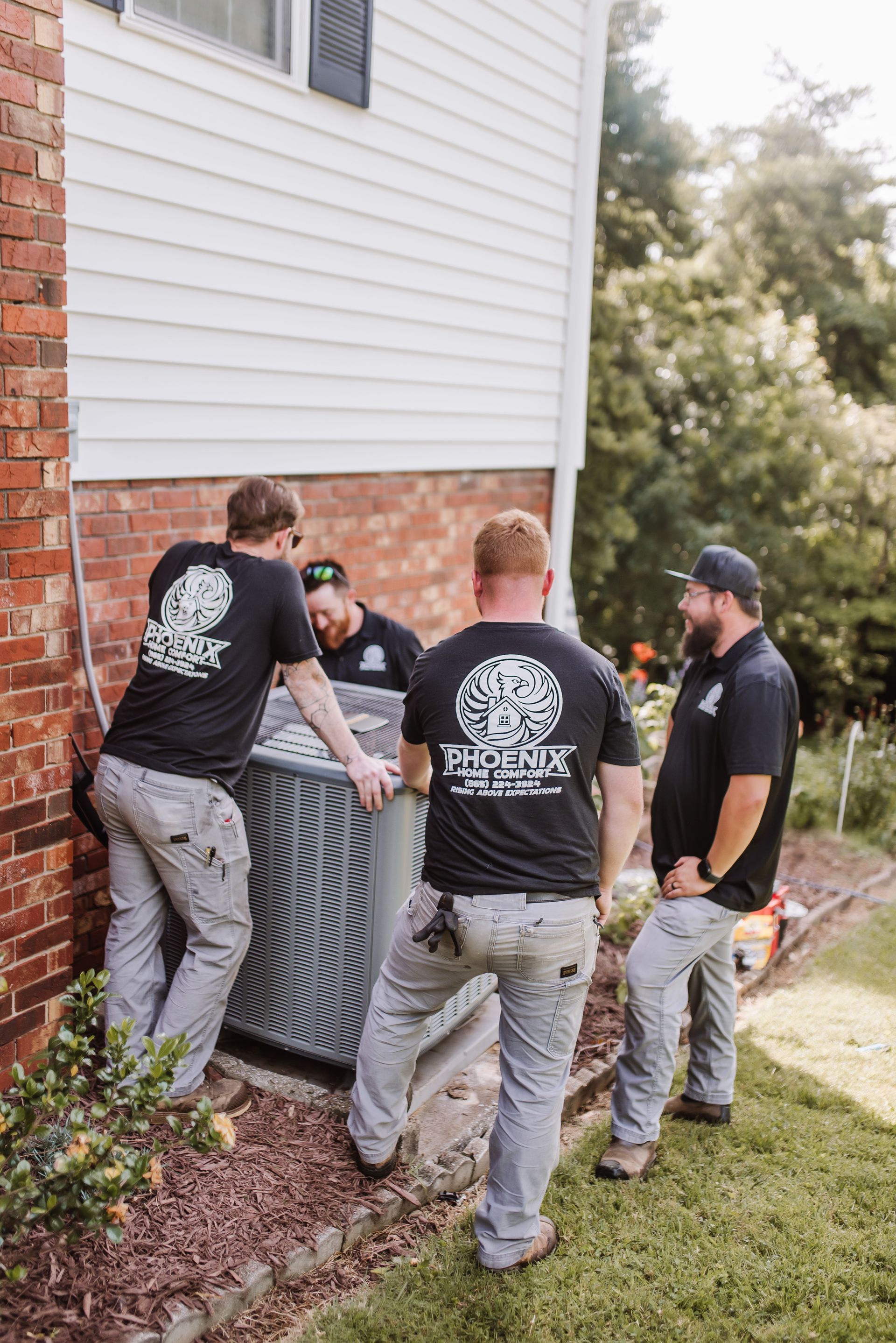 Four workers lifting an air conditioning unit near a house. They wear matching shirts and pants.