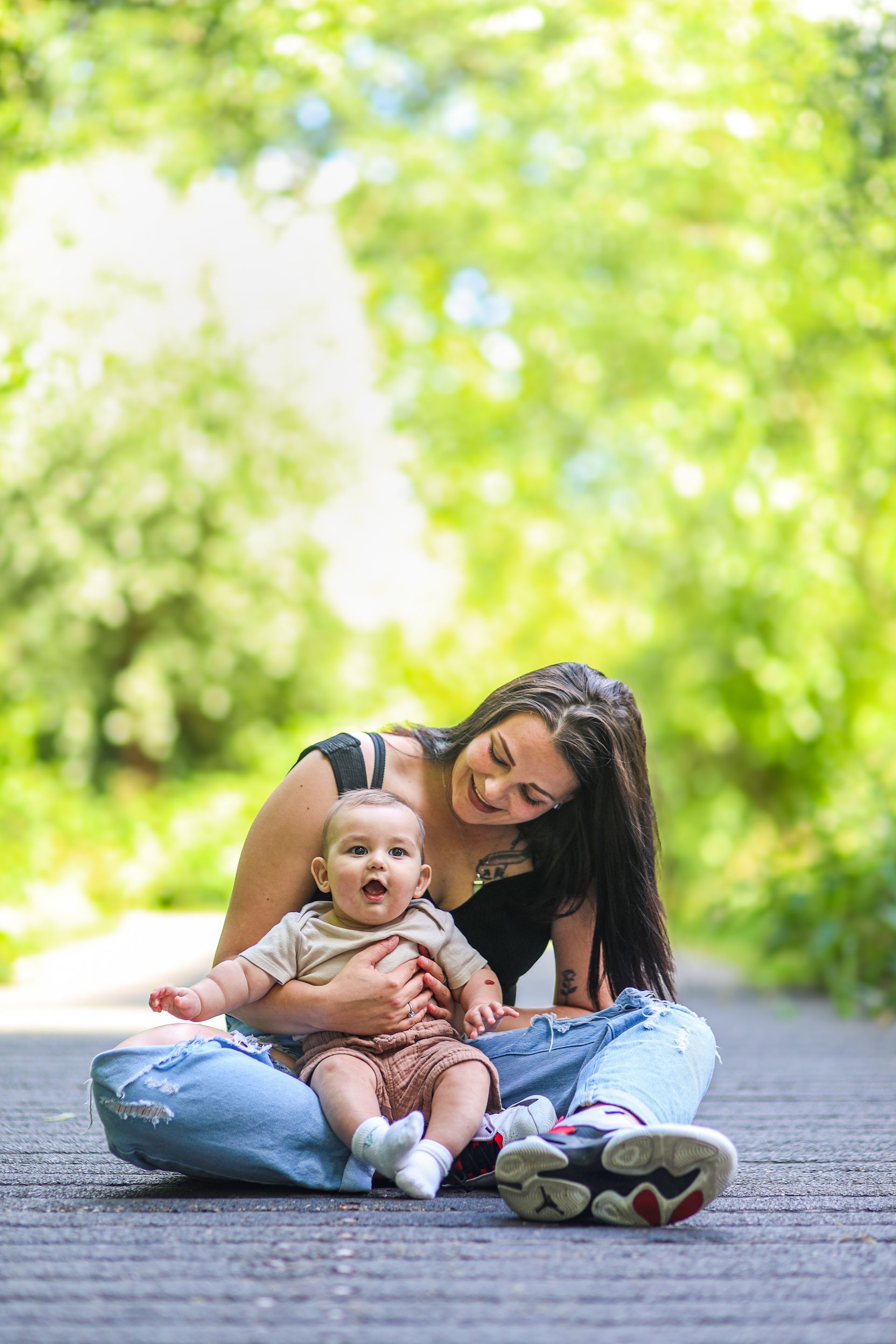 Vrouw zit op een pad met een baby op haar schoot en lacht in een zonnig park. Ouder-kind fotoshoot.