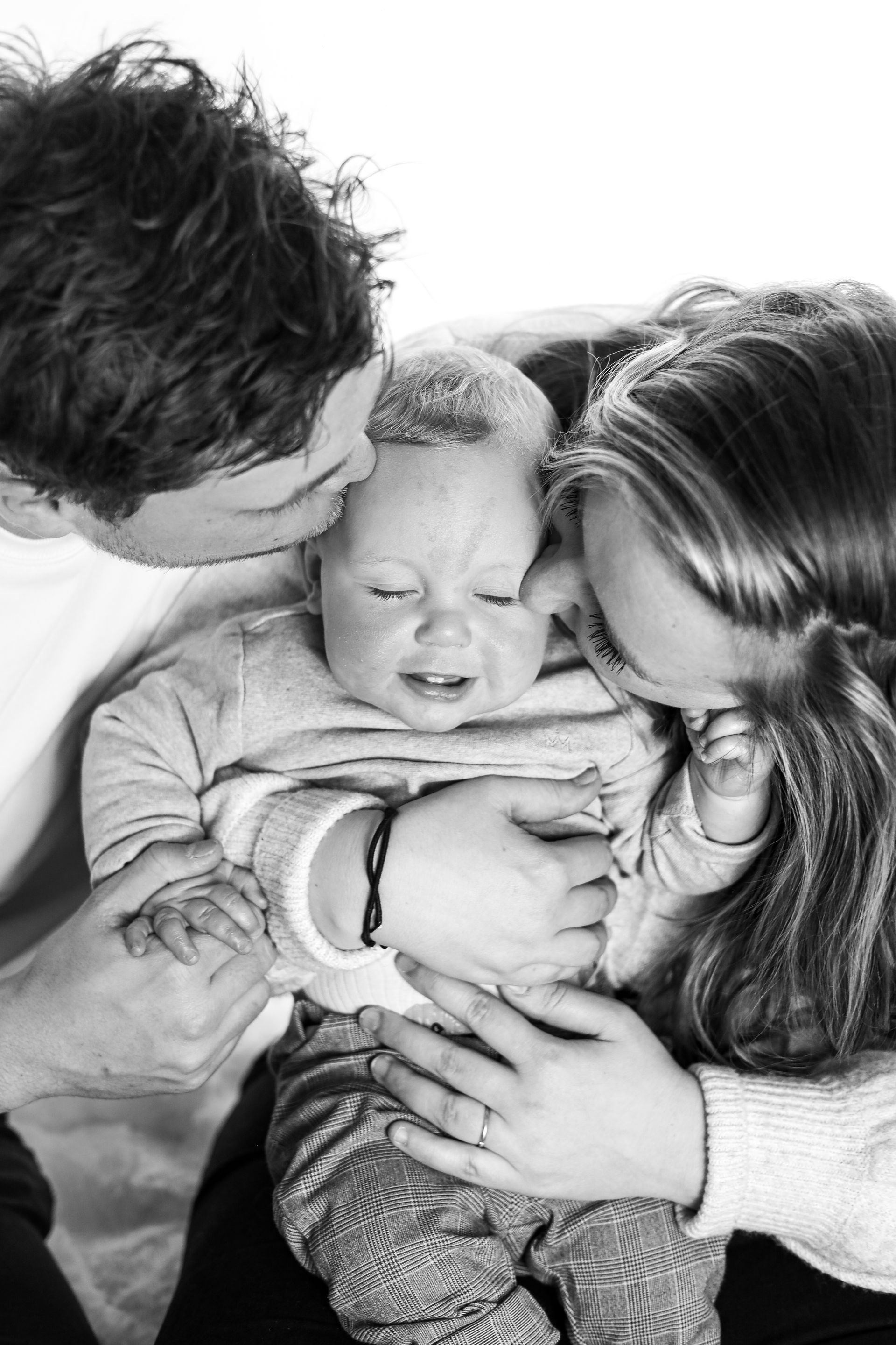 Ouders die een slapende baby kussen en dicht tegen zich aanhouden op een zwart-witfoto. Gezinsfotoshoot