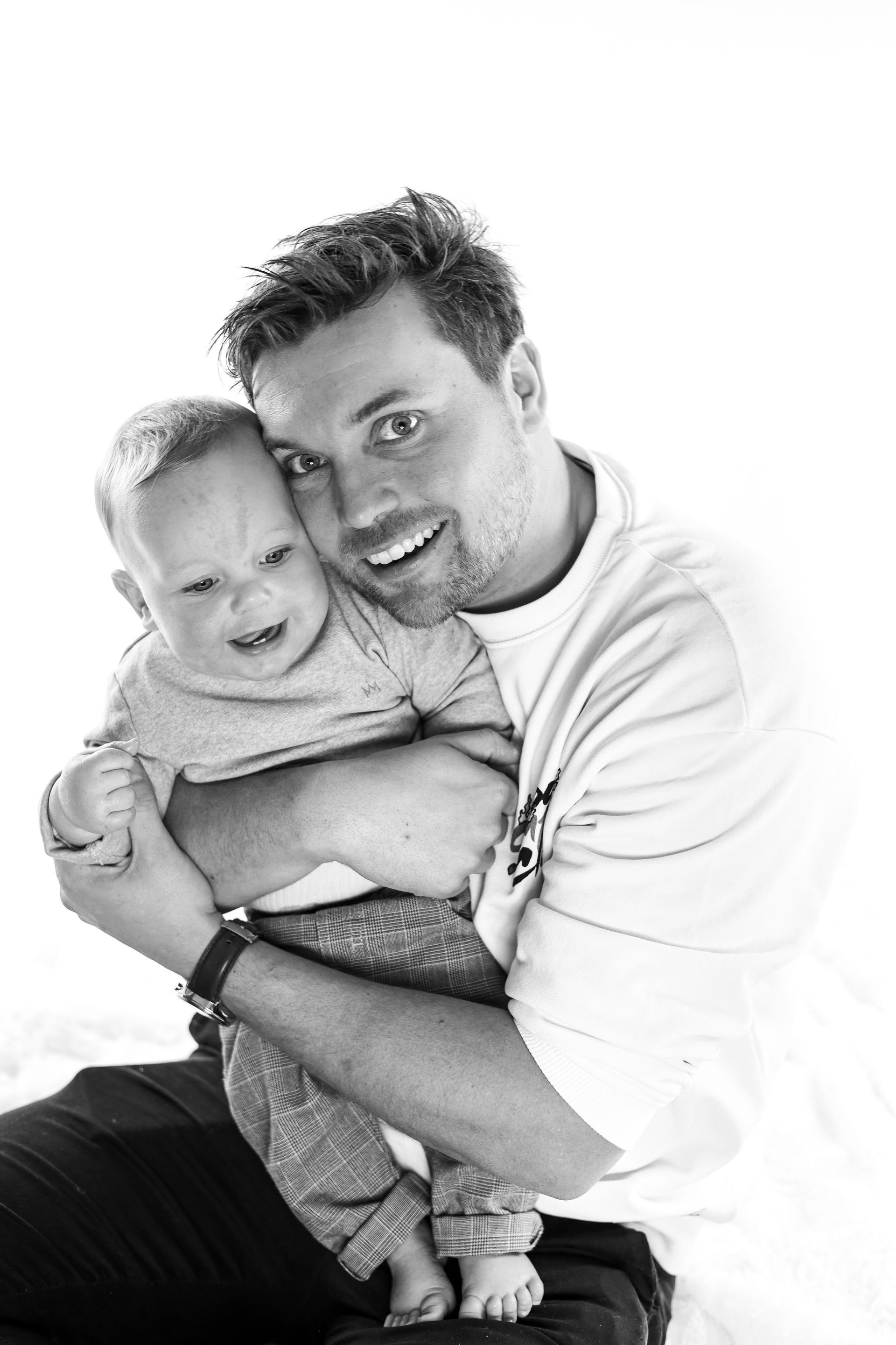 Man houdt een lachend peutertje stevig vast tegen een achtergrond van effen licht. Vader zoon fotoshoot.