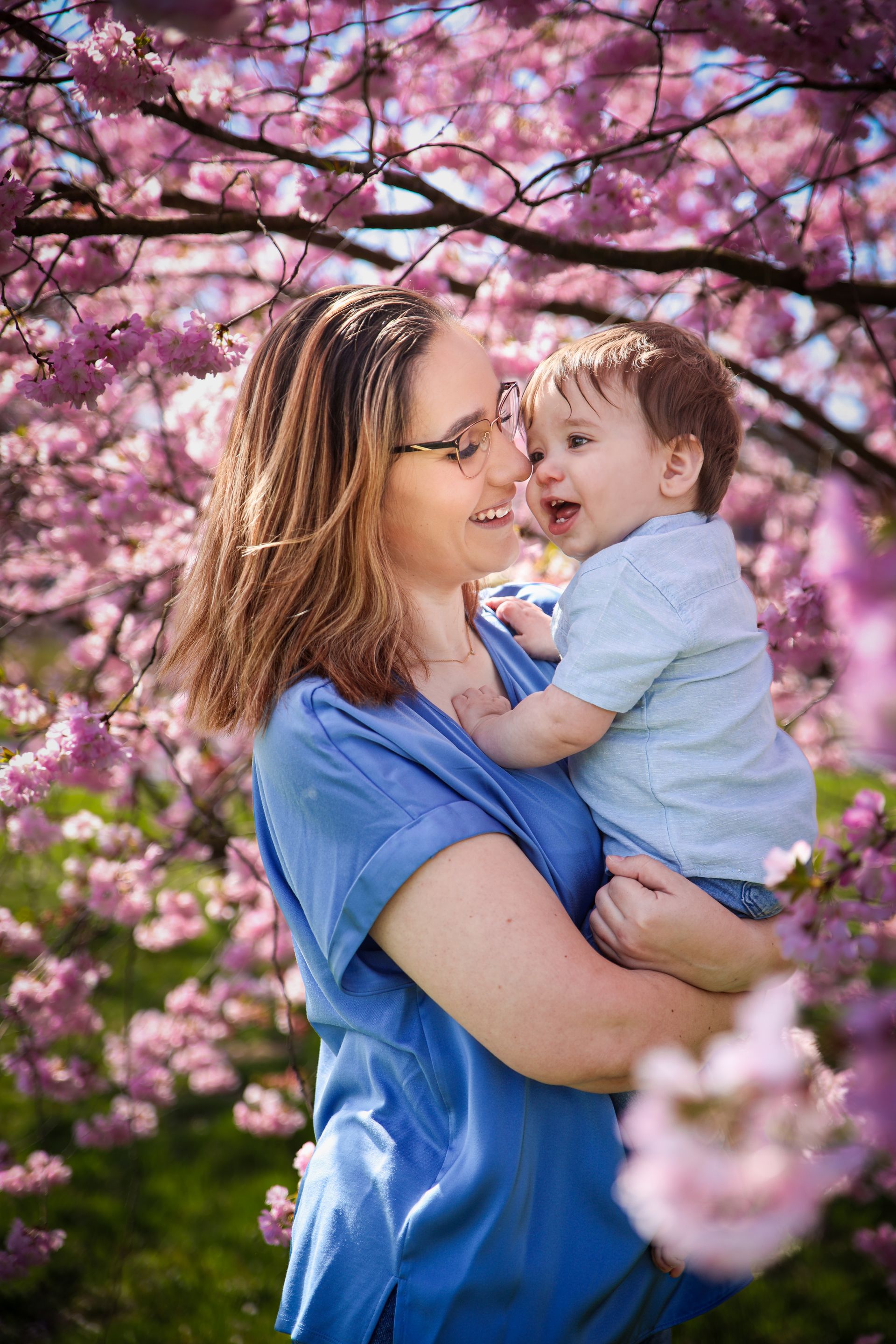 Vrouw in blauwe blouse die een lachend kind vasthoudt onder roze bloesems. Moeder dochter fotoshoot.
