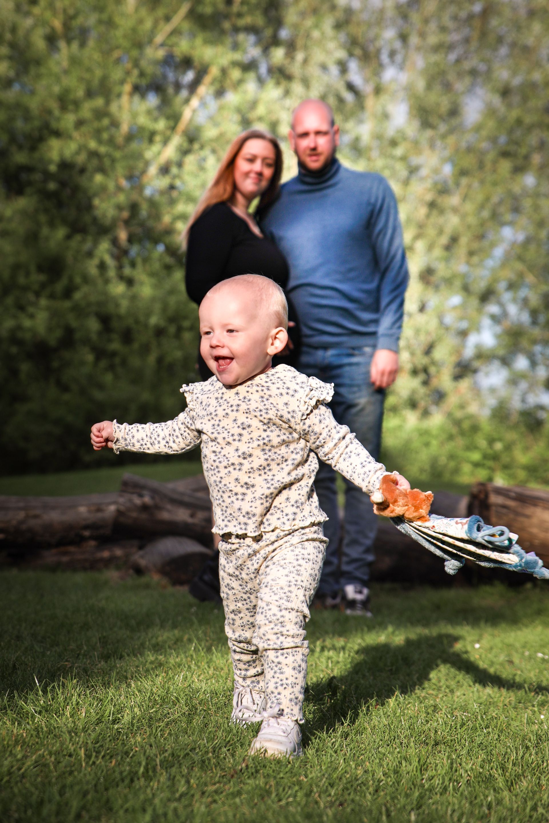 Peuter in een bedrukte romper loopt over het gras met twee volwassenen erachter in een zonnig park. Genderreveal fotoshoot.