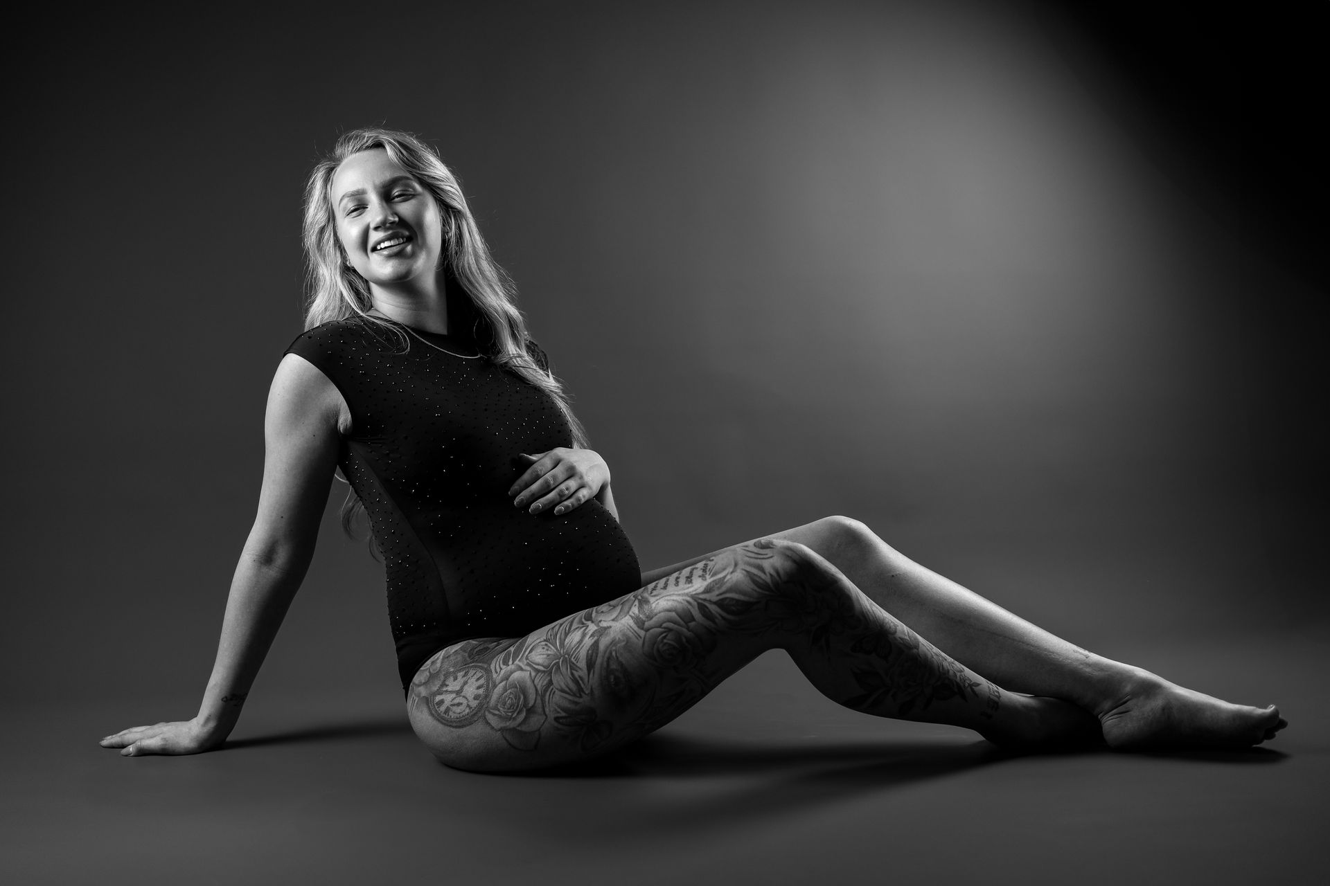 Vrouw ligt in een zwarte outfit op de grond en lacht in een studio met een donkere achtergrond. Zwangerschap fotografie.