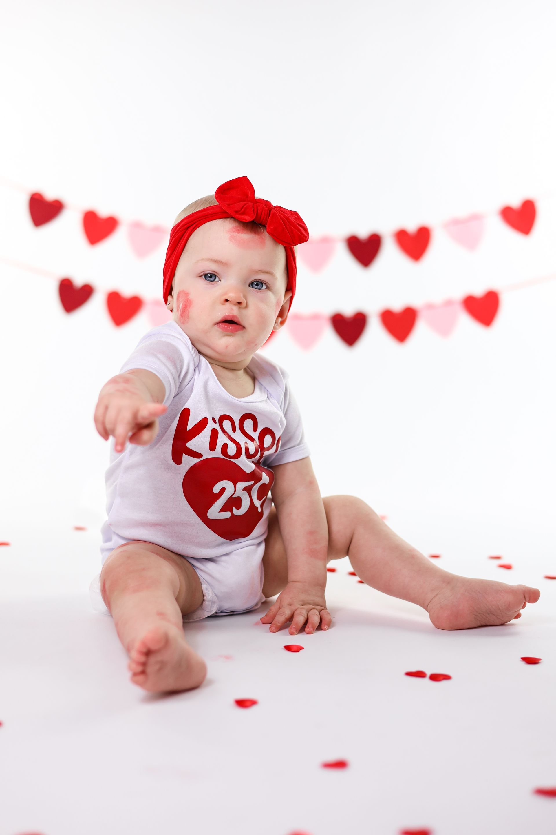 Een baby in een wit rompertje met een rode strik zit op een witte achtergrond met rode hartjes. Valentijn fotoshoot.