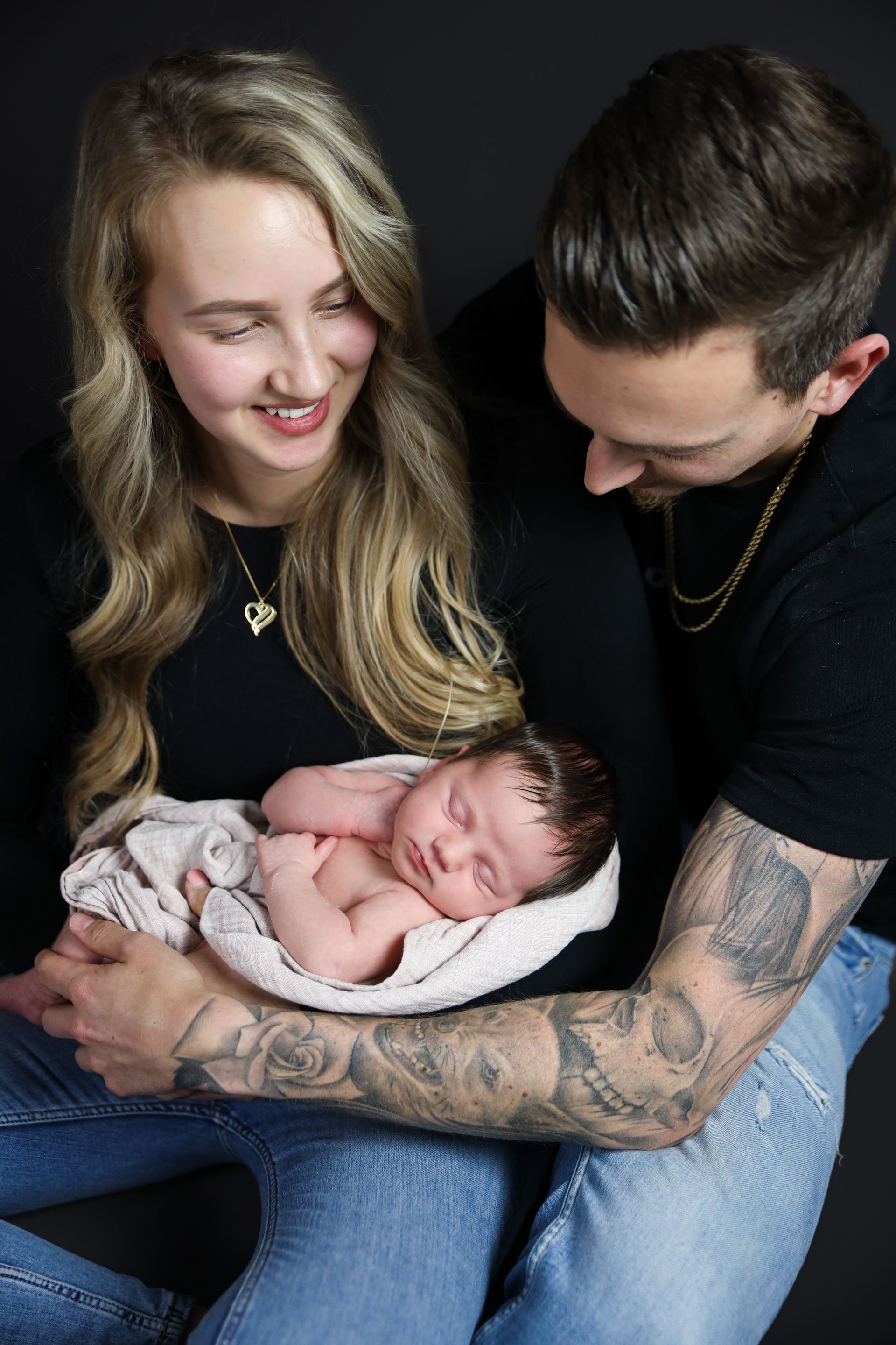 Een echtpaar houdt een slapende pasgeborene op schoot en kijkt er teder op neer. Newborn fotograaf.