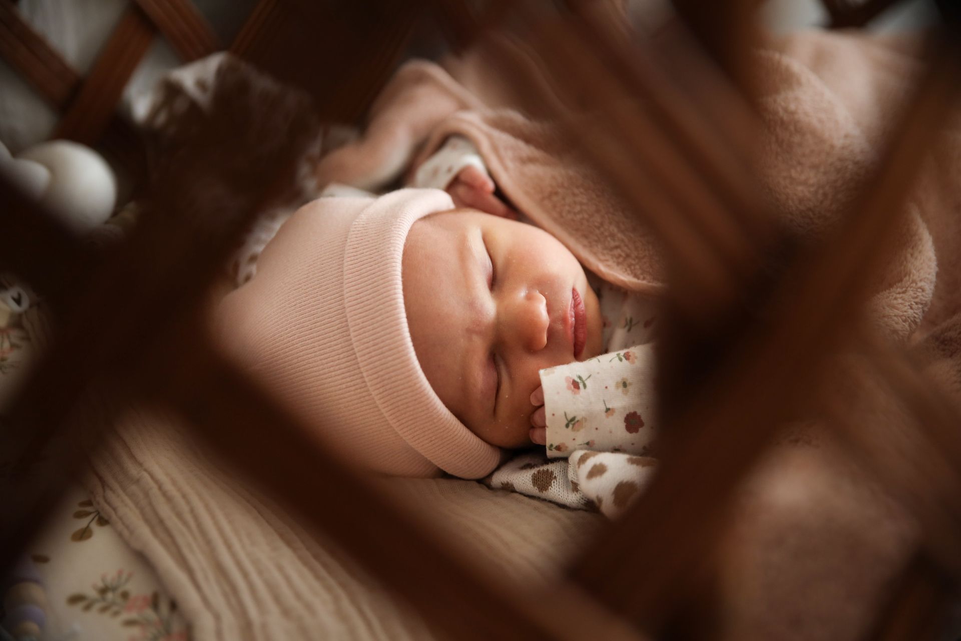 Een slapende baby met een roze gebreid mutsje is te zien door de wazige spijlen van het babybedje. Newborn fotografie.